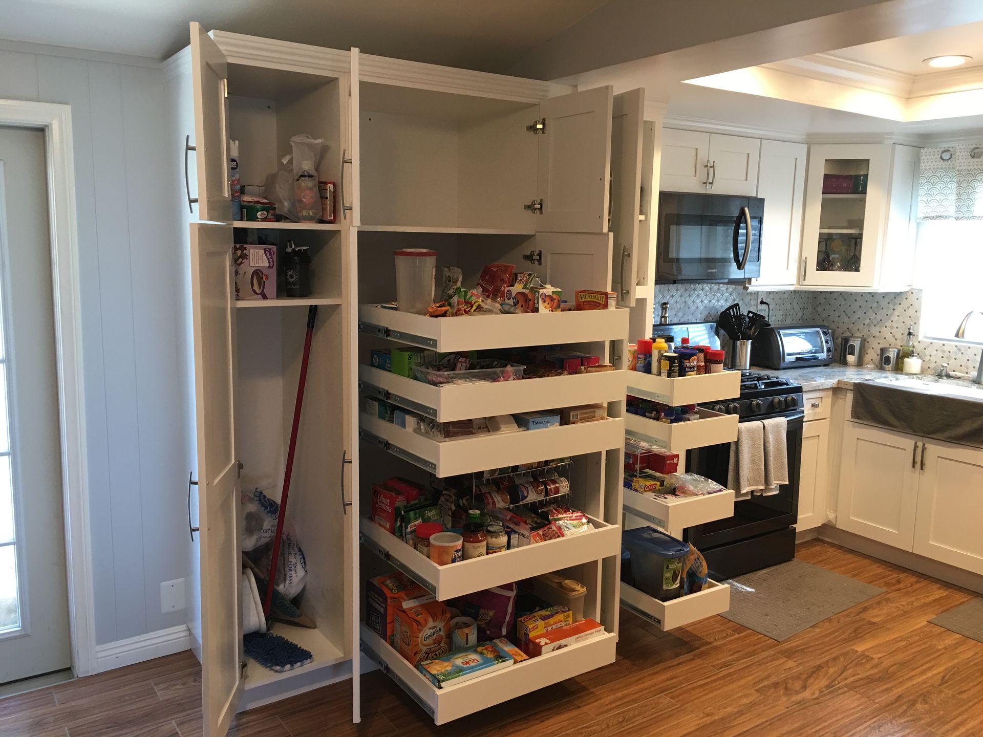 White kitchen pantry with pull-out shelves filled with food items. Broom and cleaning supplies in side cabinet.