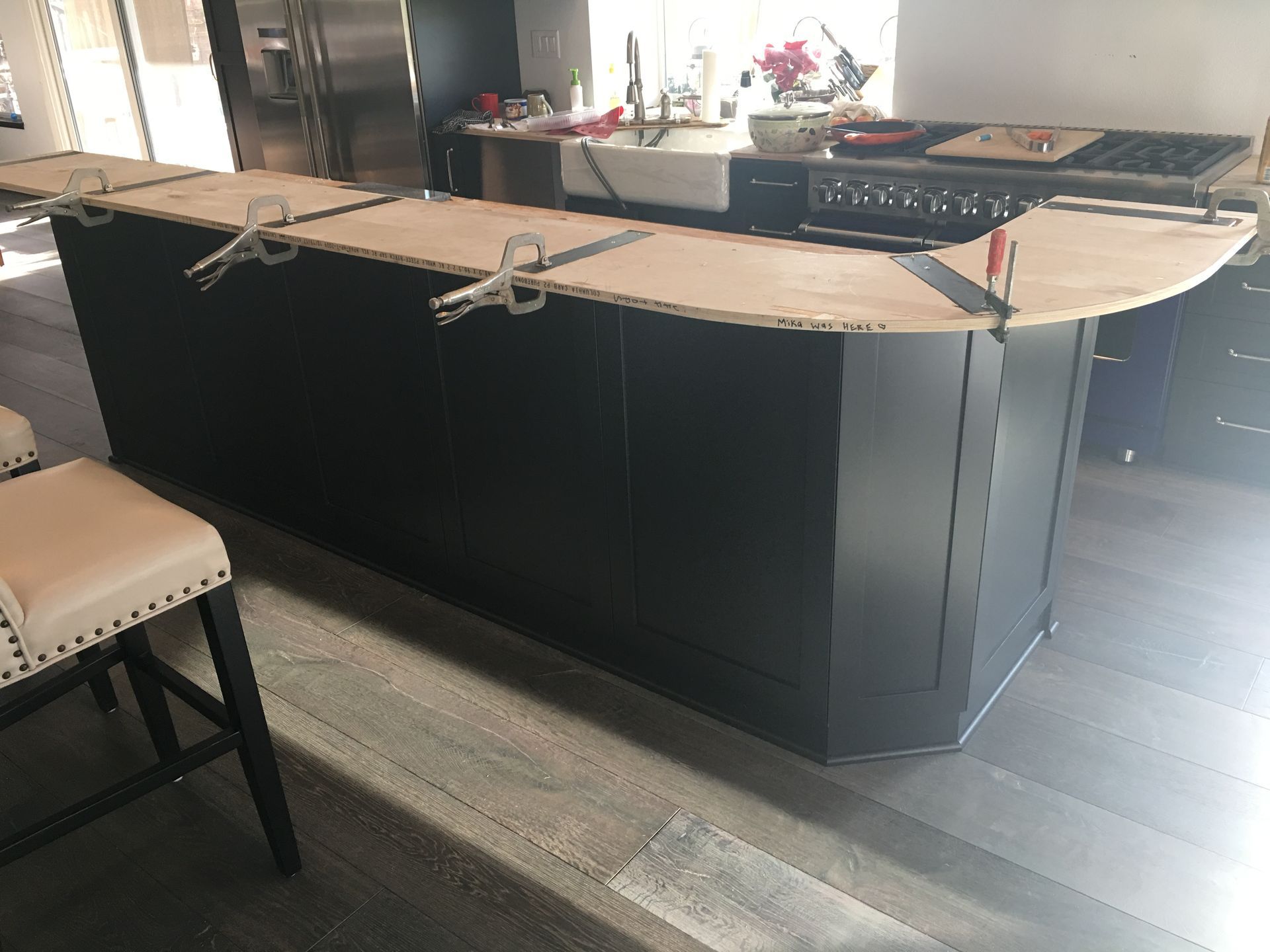 Black kitchen island under construction with clamped plywood countertop; stools in foreground.