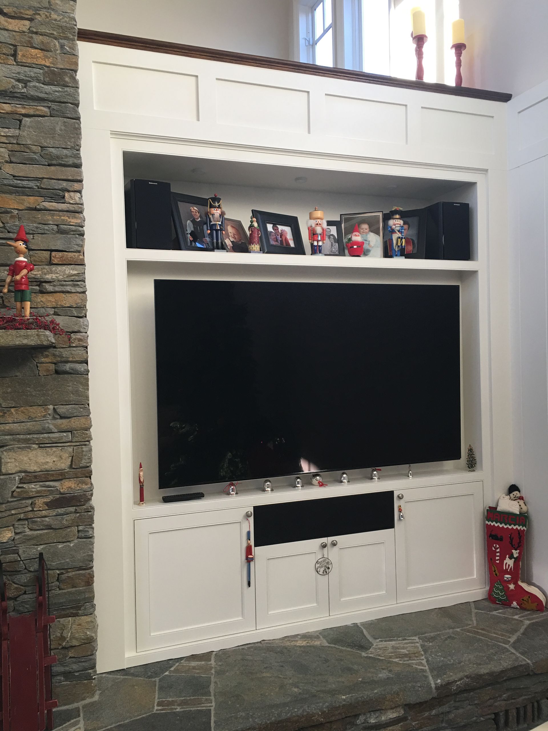 Built-in white media center with TV, upper shelf with decor, and cabinets on a stone hearth.
