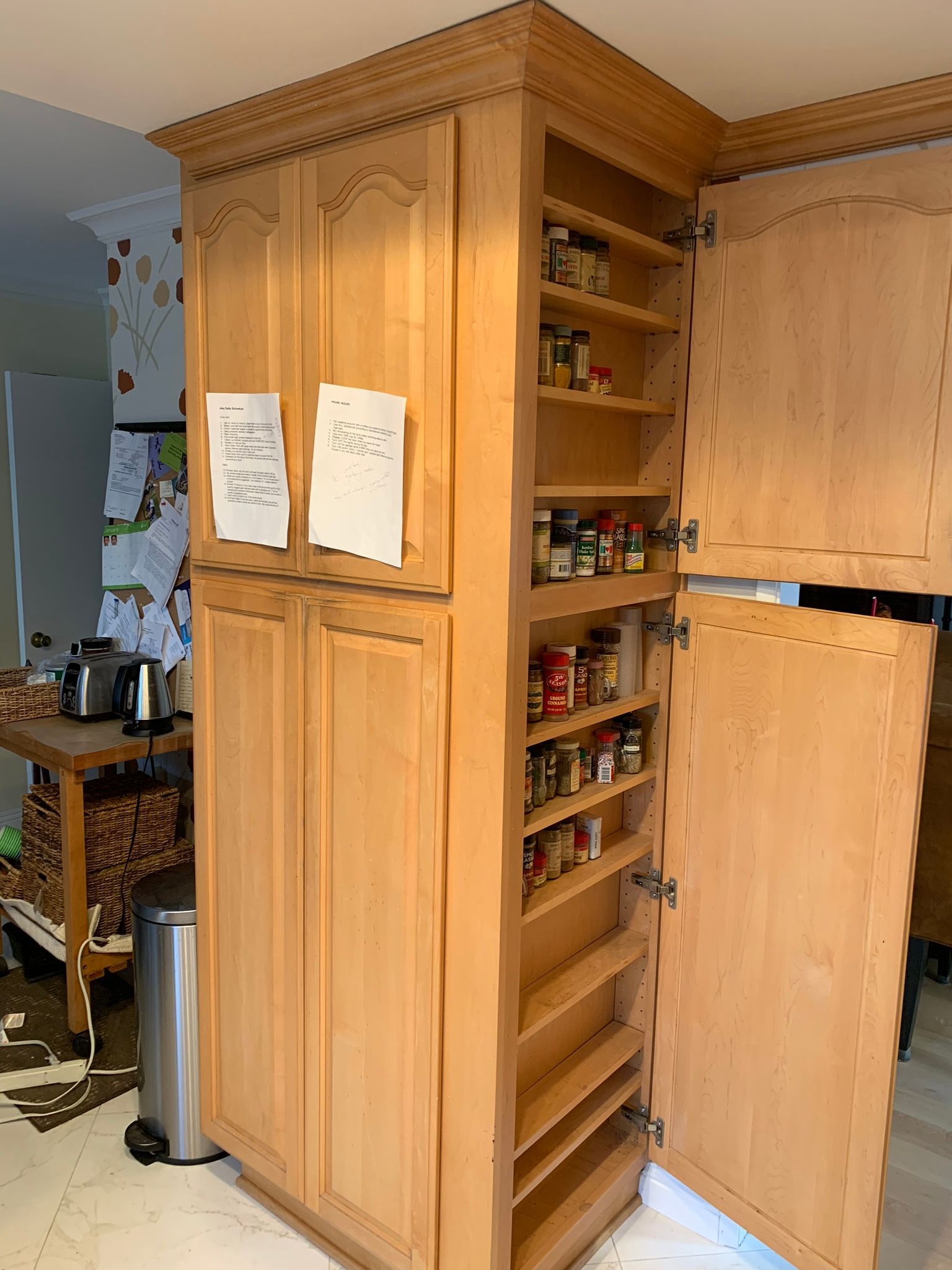 Tall wooden pantry cabinet with open spice rack, located in a kitchen.