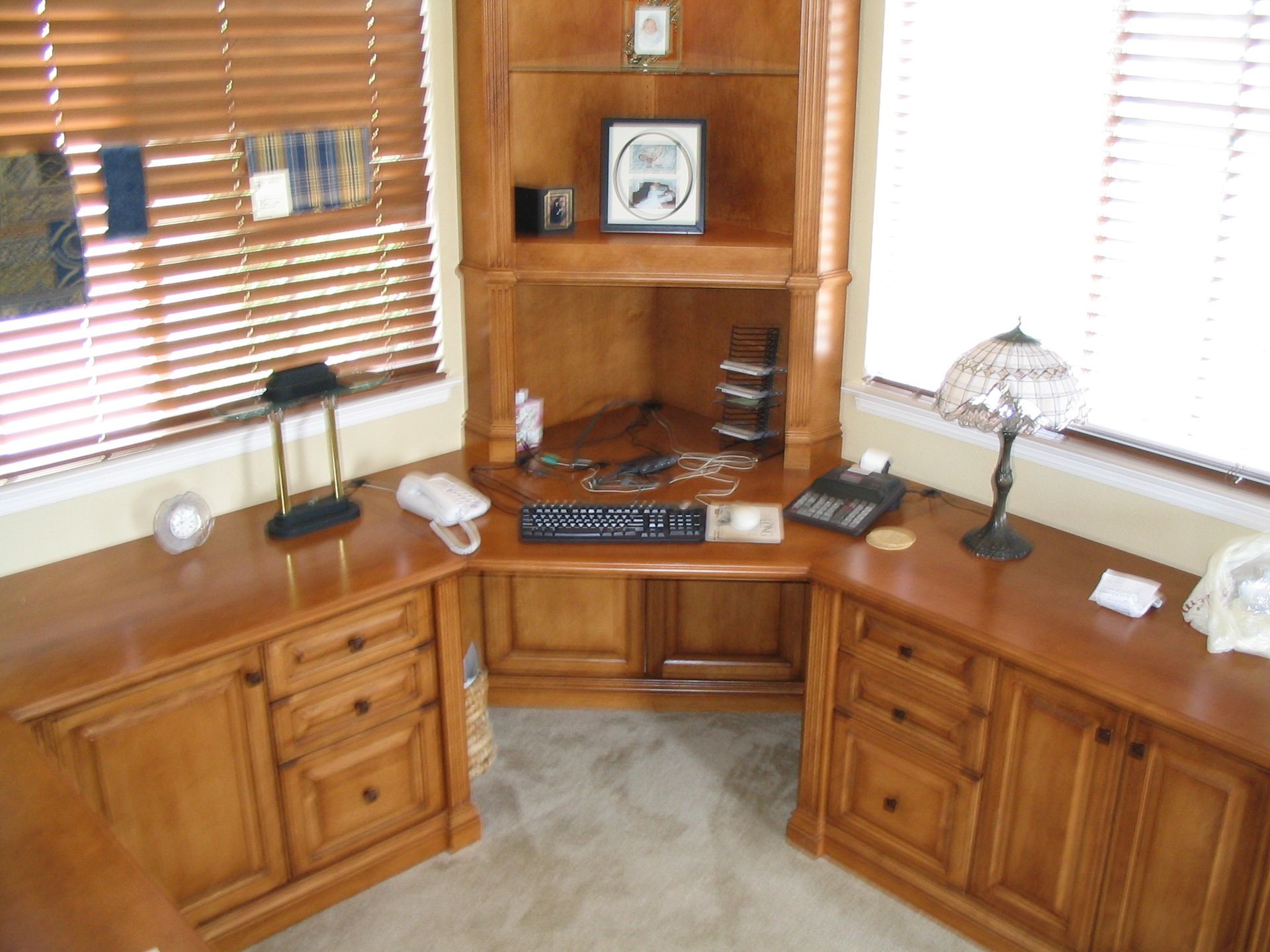 Wooden corner desk with built-in shelves and drawers, near a window with blinds.