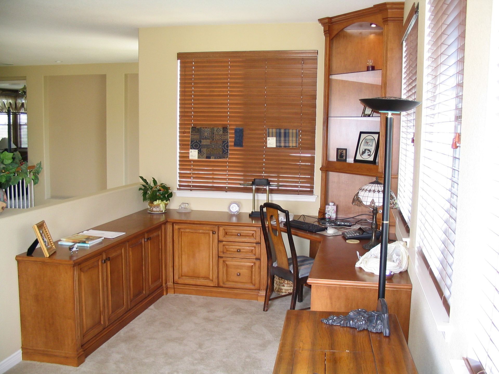 Wooden home office desk with cabinets and shelving near a window with blinds.