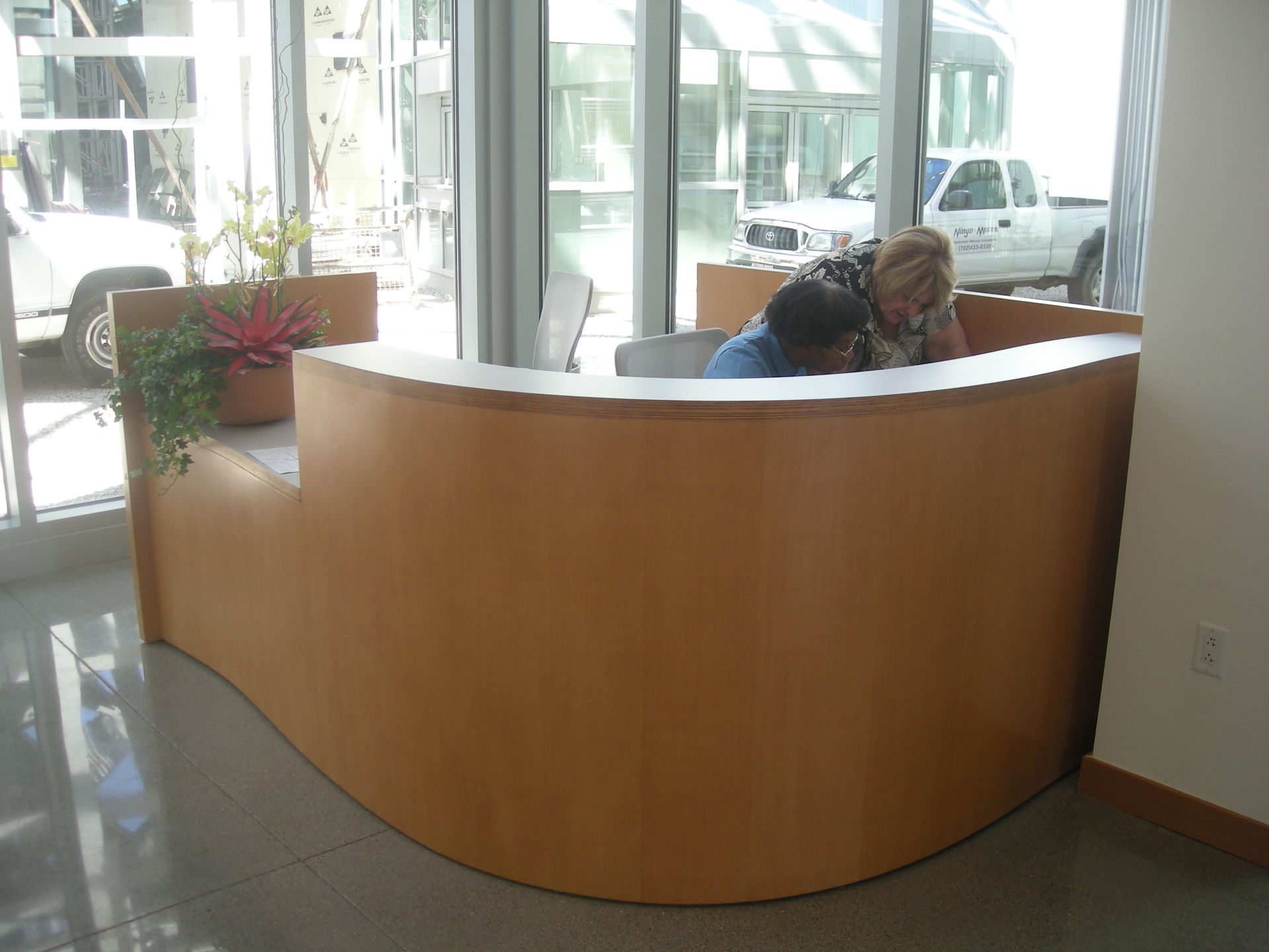 Curved wooden reception desk; two people interact, sunny interior.