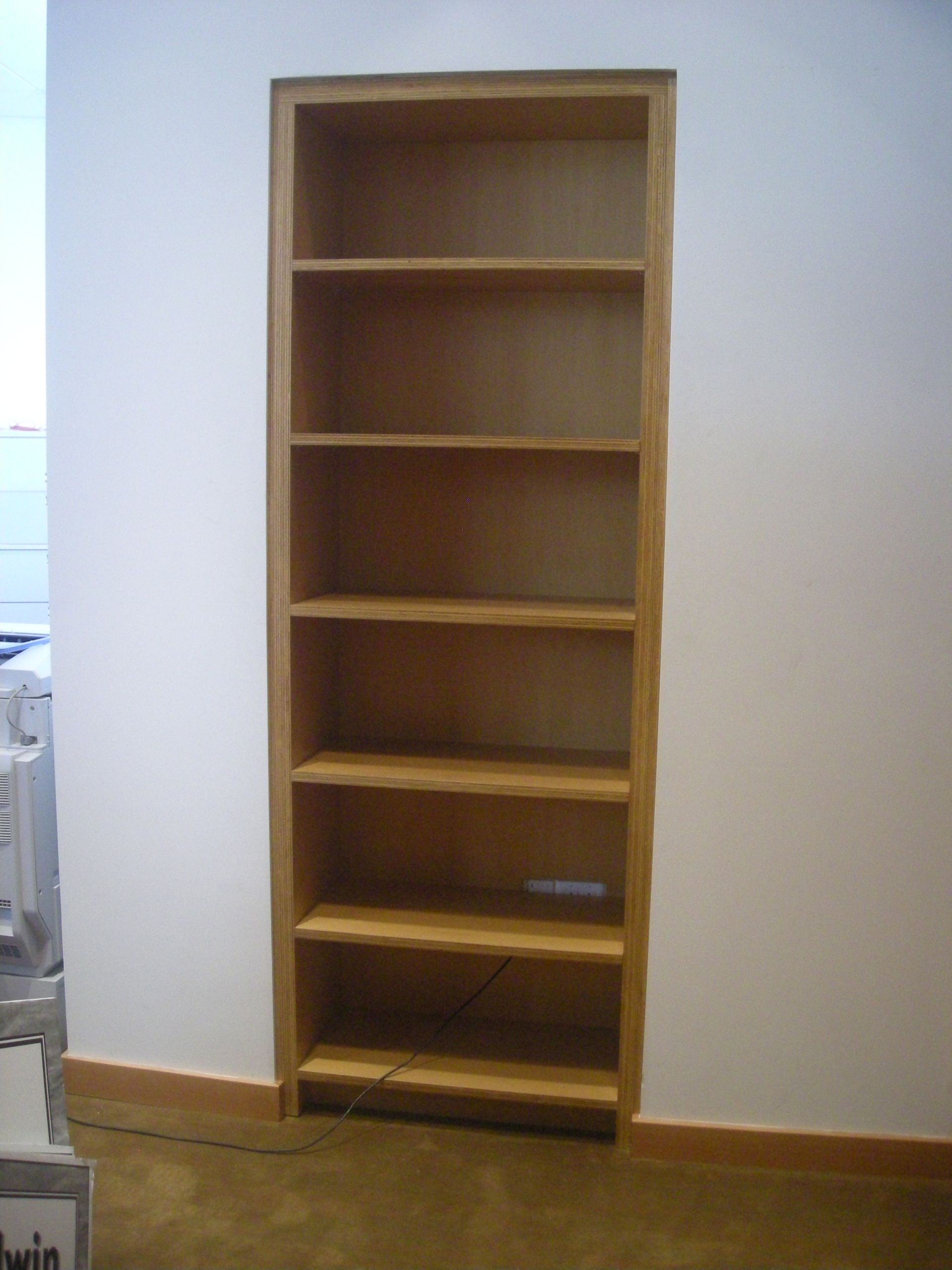 Wooden built-in bookshelf with six shelves in a white-walled room, set against a brown baseboard.