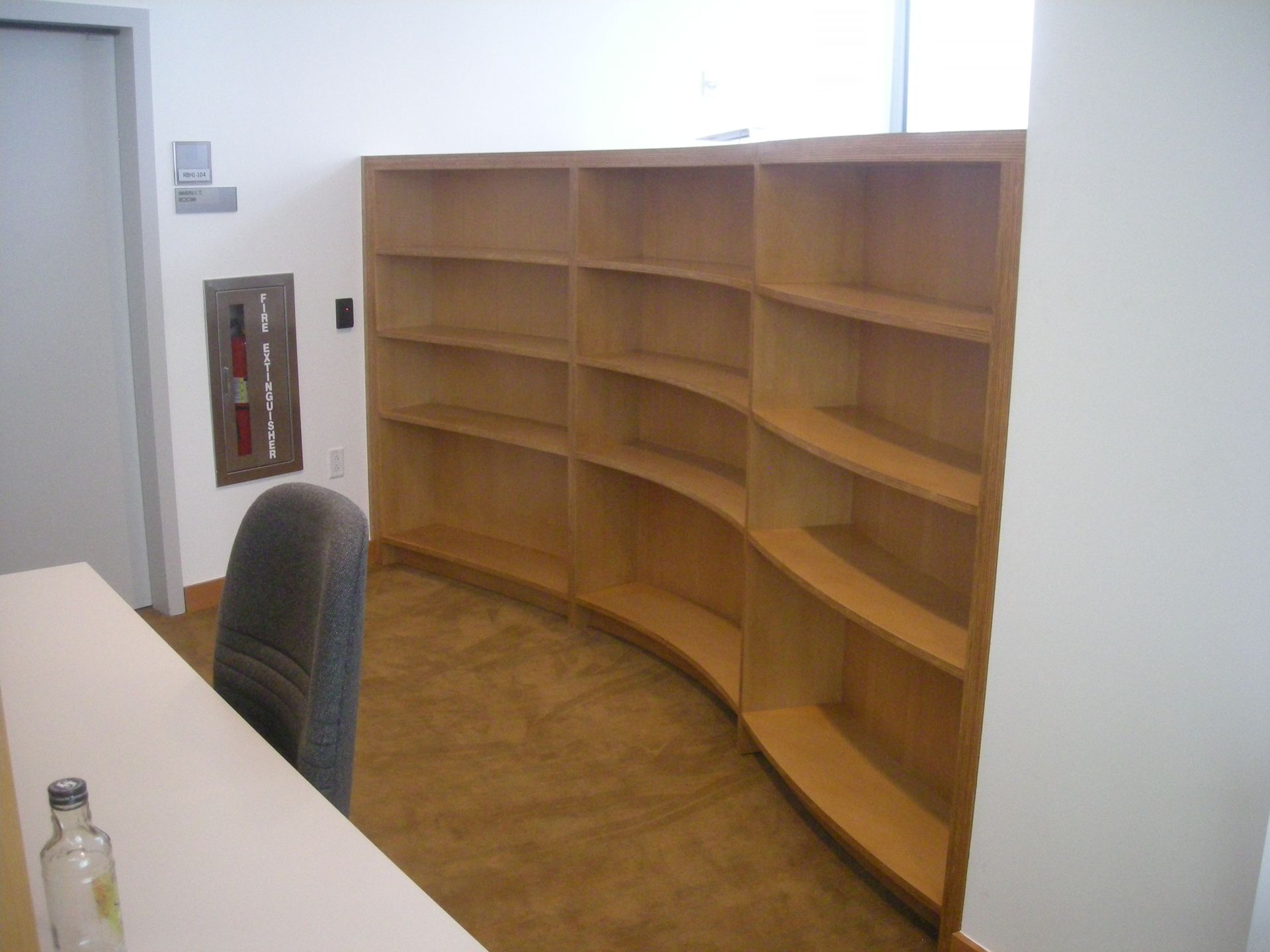 Empty curved wooden bookshelves in a corner of a room, next to a chair and a fire extinguisher.