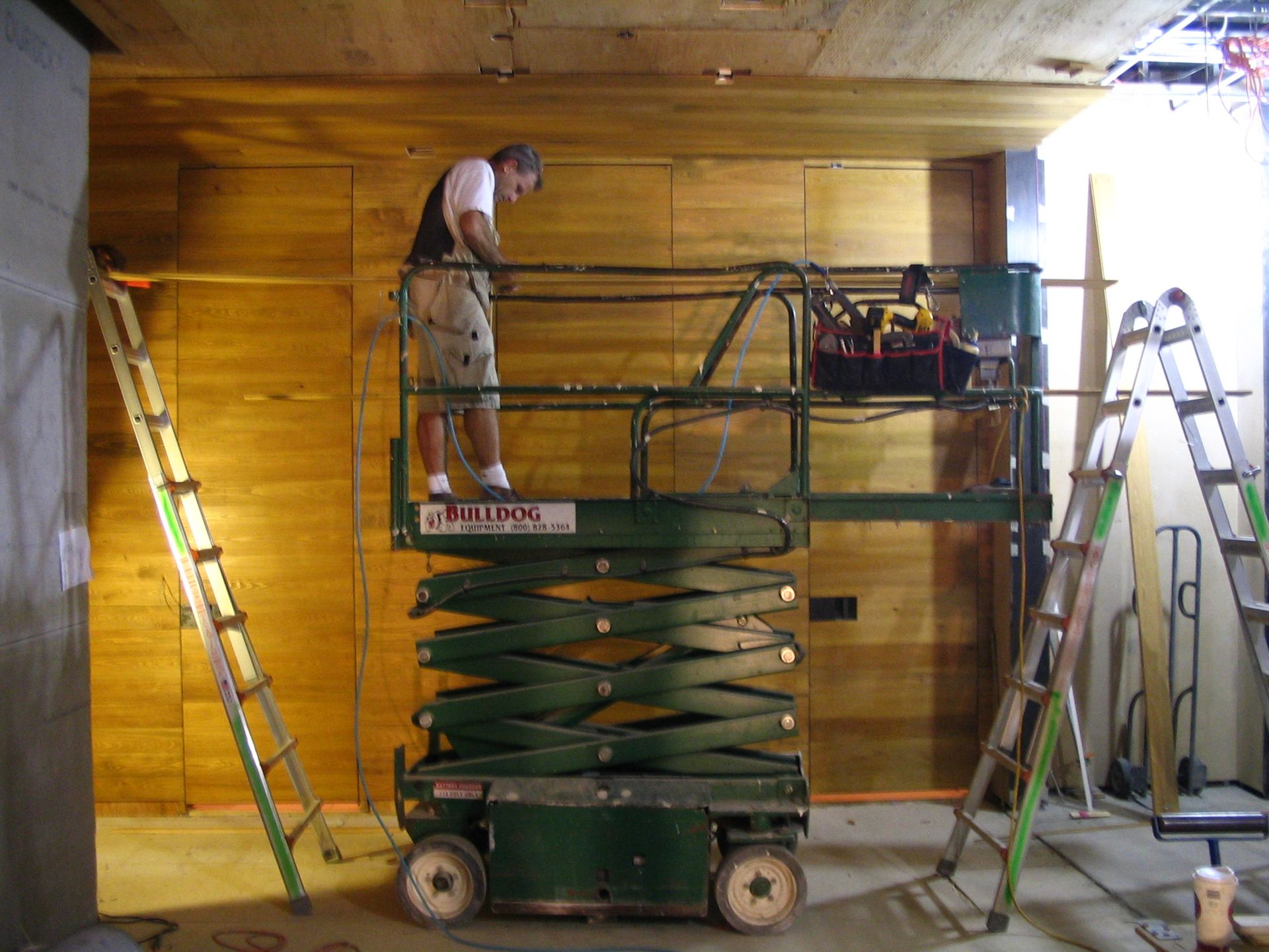 Man on lift working on wood paneling, surrounded by ladders in a construction setting.