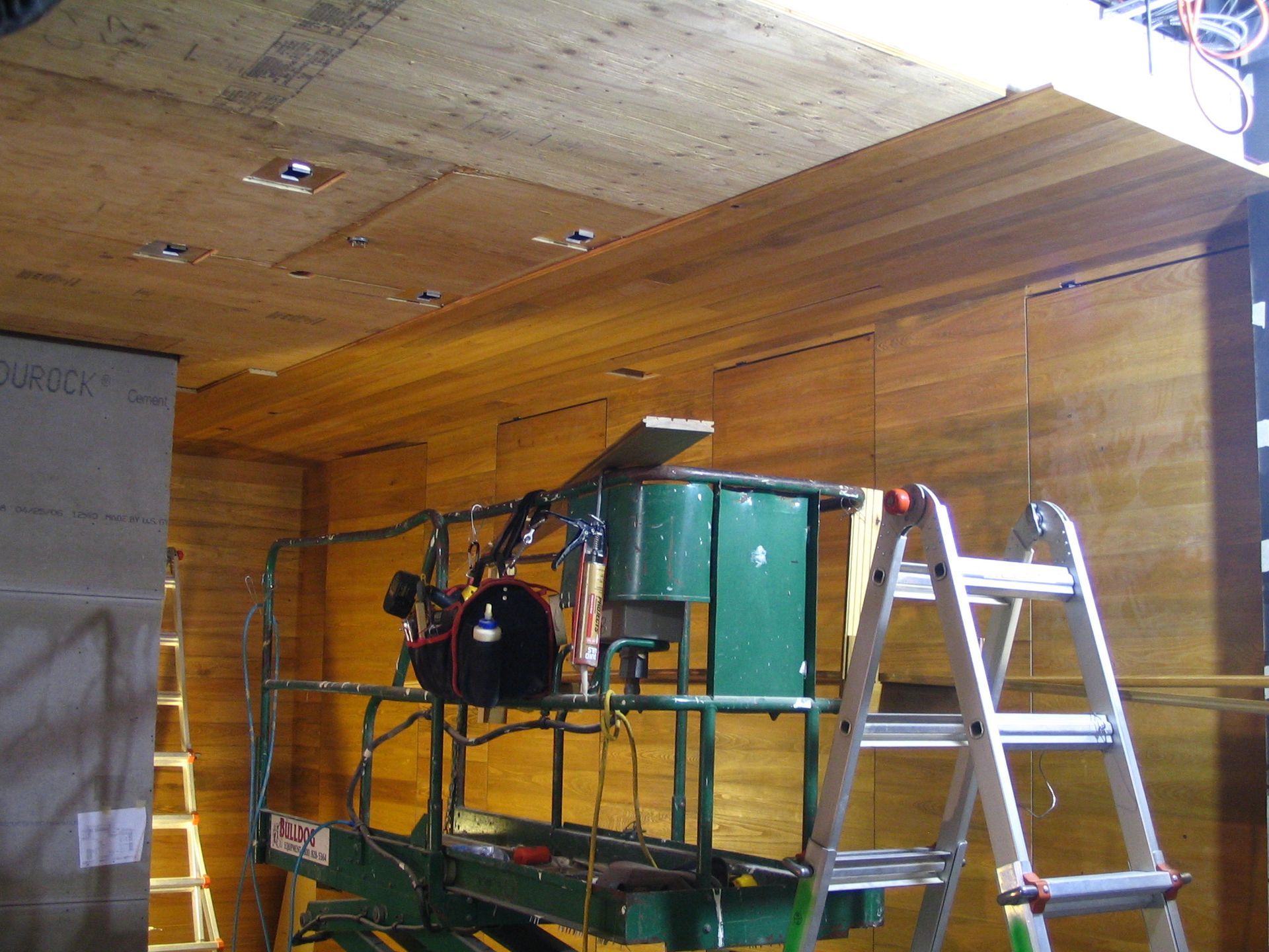 Interior construction with plywood walls and ceiling, work platform, ladder, and lights.
