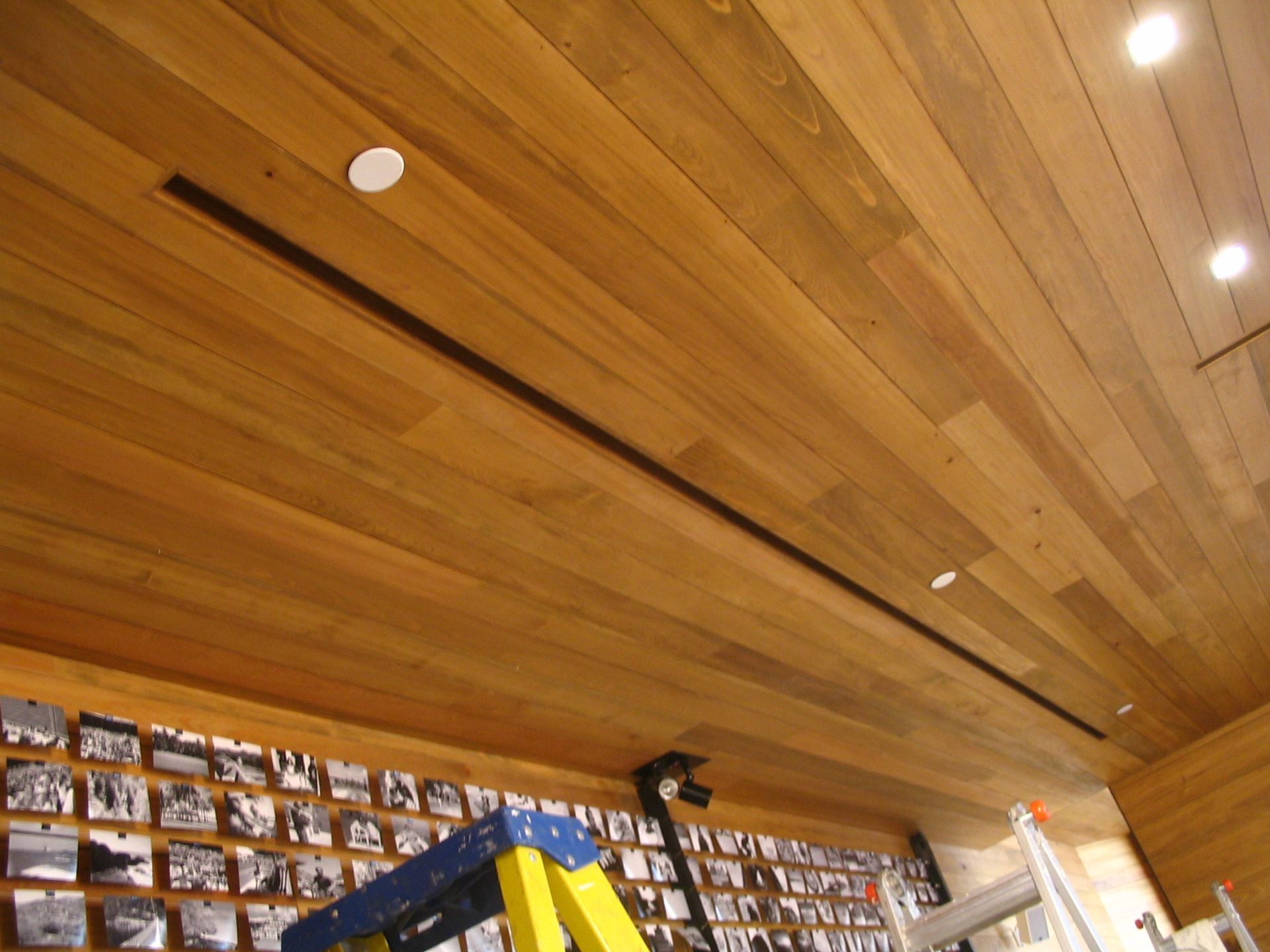 Wooden ceiling with linear black vent and recessed lights. Framed black and white photos below.