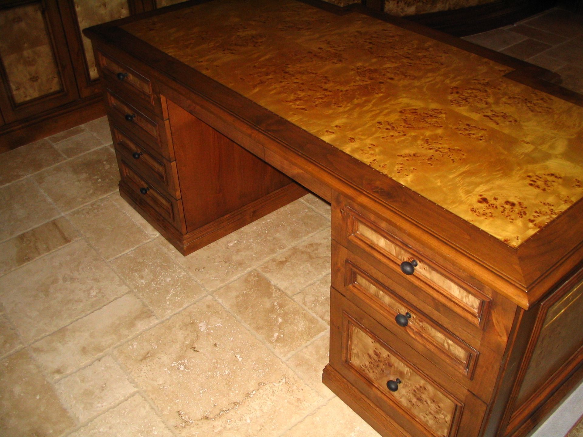 Wooden desk with drawers, ornate burl wood top, and a tile floor setting.