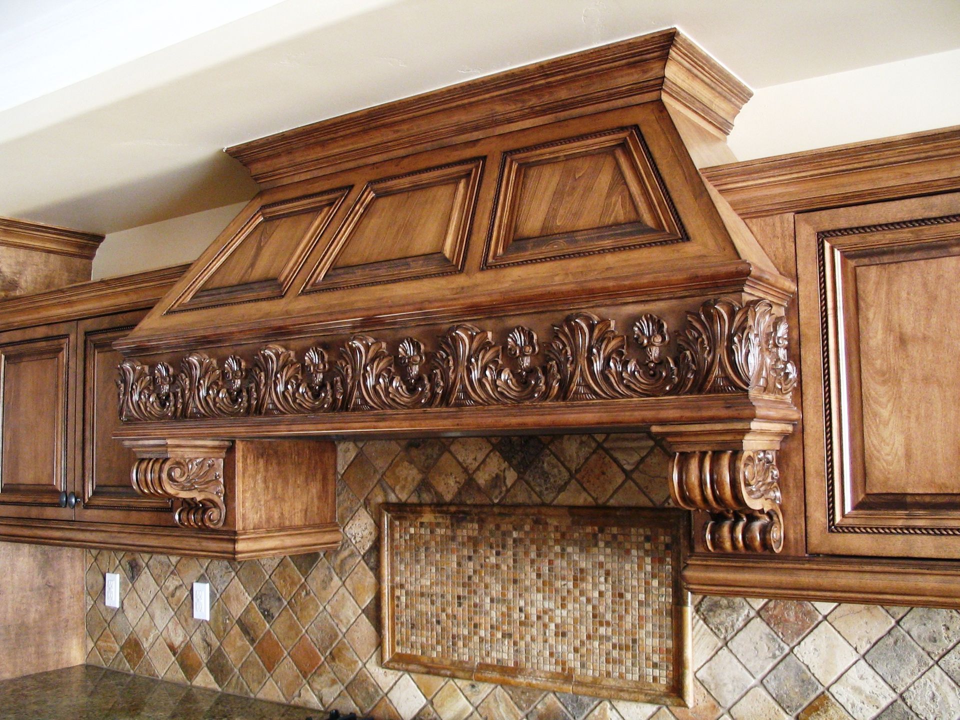 Ornate, carved wood kitchen range hood above tile backsplash and countertops.