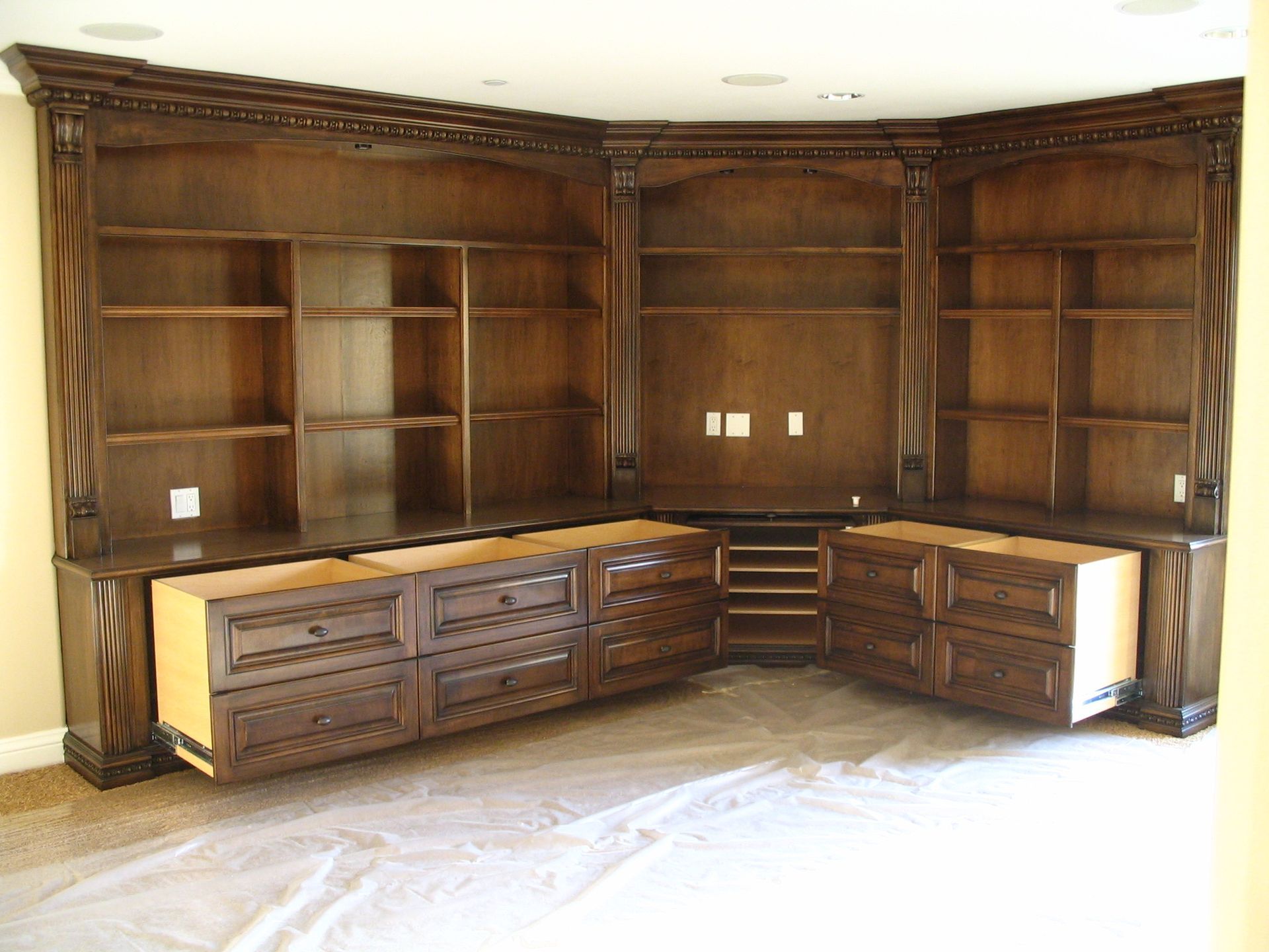 Dark wood built-in shelving and cabinets in a corner, with drawers pulled out. Empty room, tan walls.