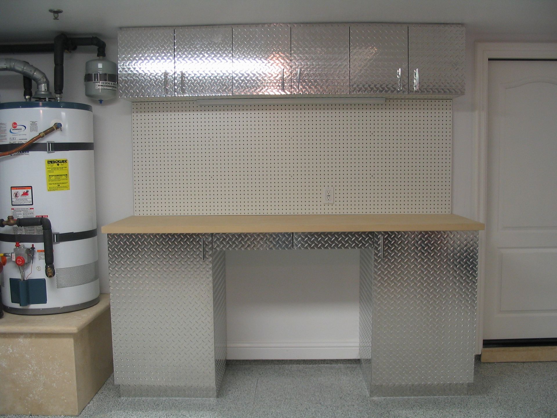 Garage workspace with diamond plate metal cabinets, pegboard, and workbench. Water heater visible on the left.