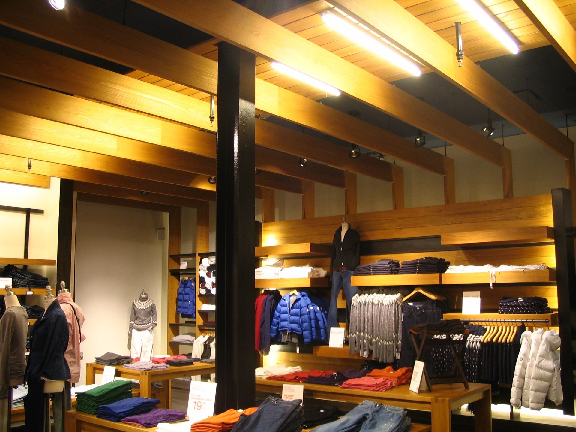 Clothing store interior with wood beams, displaying clothes on shelves and tables under bright lights.
