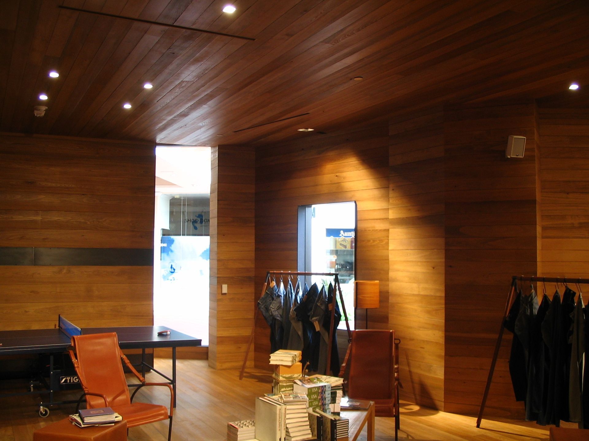 Wooden interior of a clothing store with orange chairs, racks of clothes, and a bright doorway.