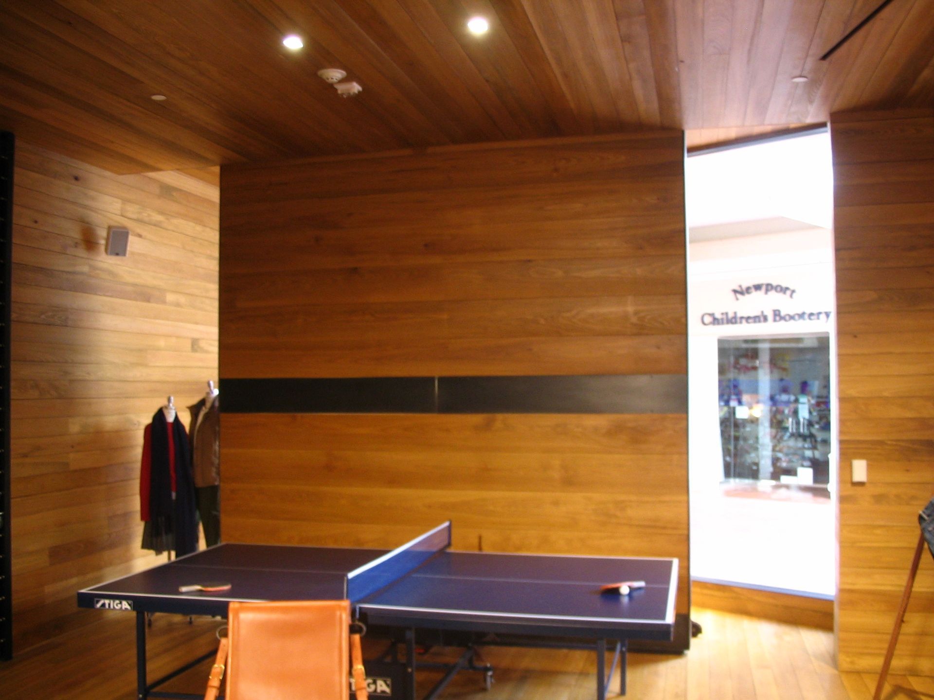 Ping pong table in wood-paneled room, opening to a store with glass doors.