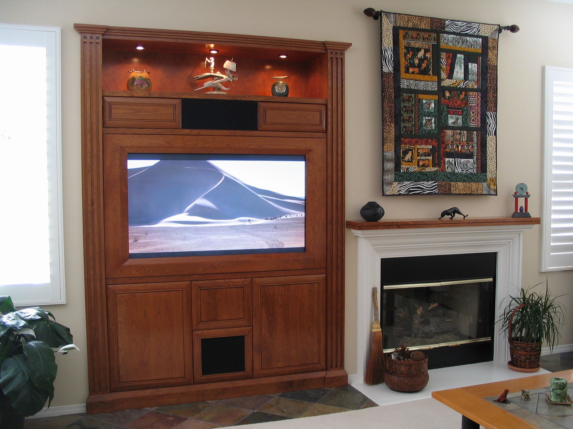Wooden media center with a TV, above a fireplace with a decorative quilt hanging on the wall.