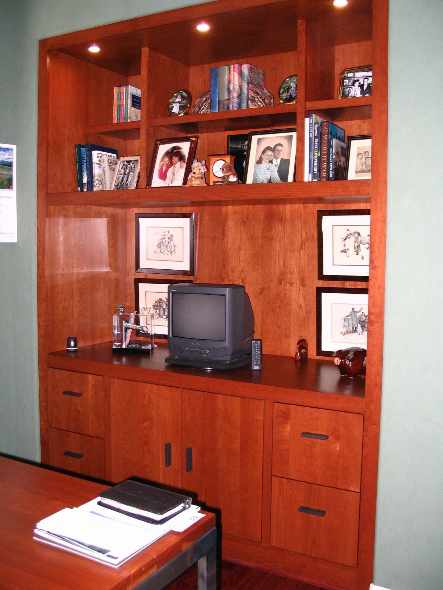 Built-in cherry wood home office with shelves, drawers, desk area, and computer monitor; lit by overhead lights.