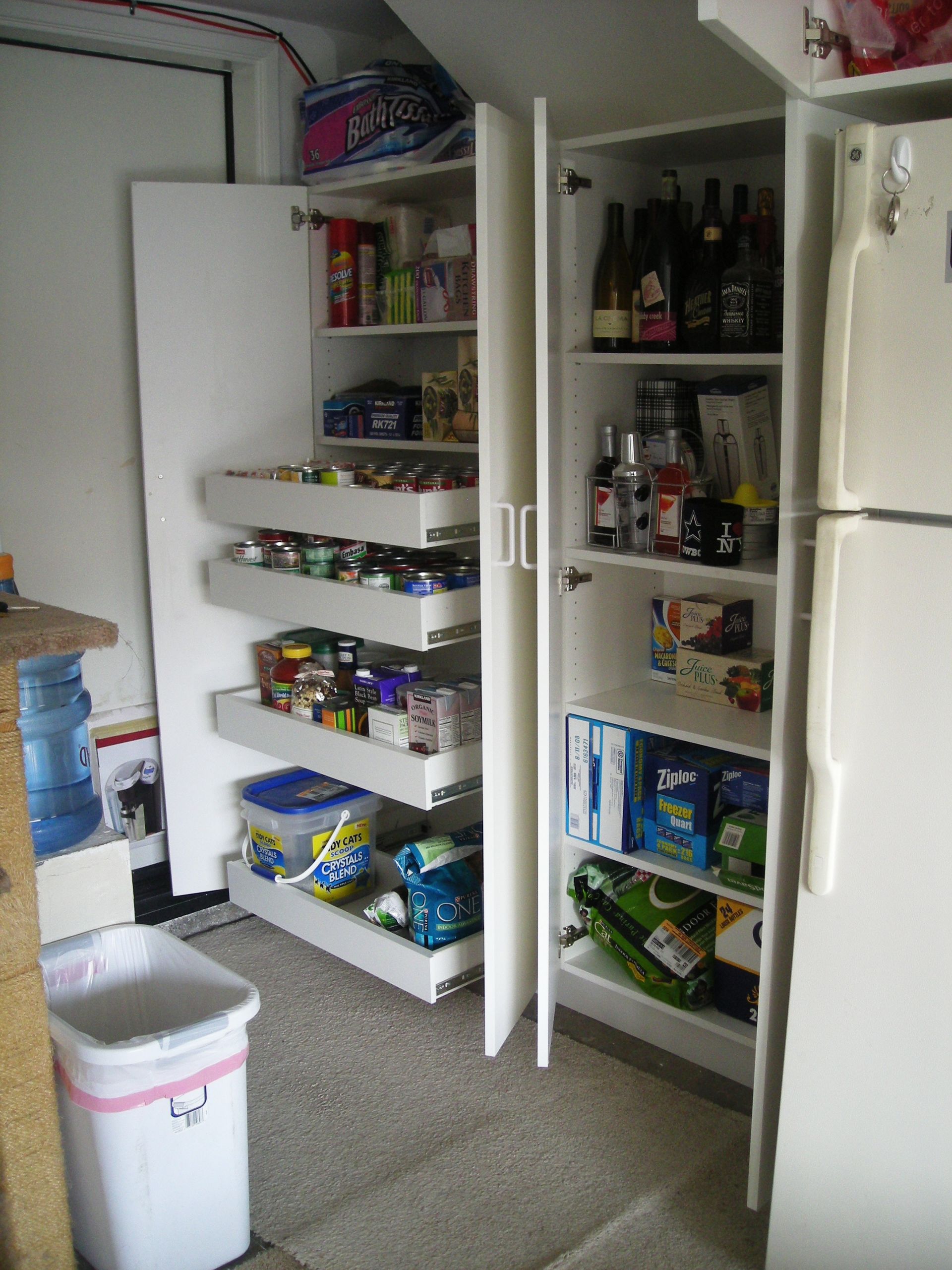 White pantry cabinets with pull-out shelves, storing food items next to a refrigerator and trash can.
