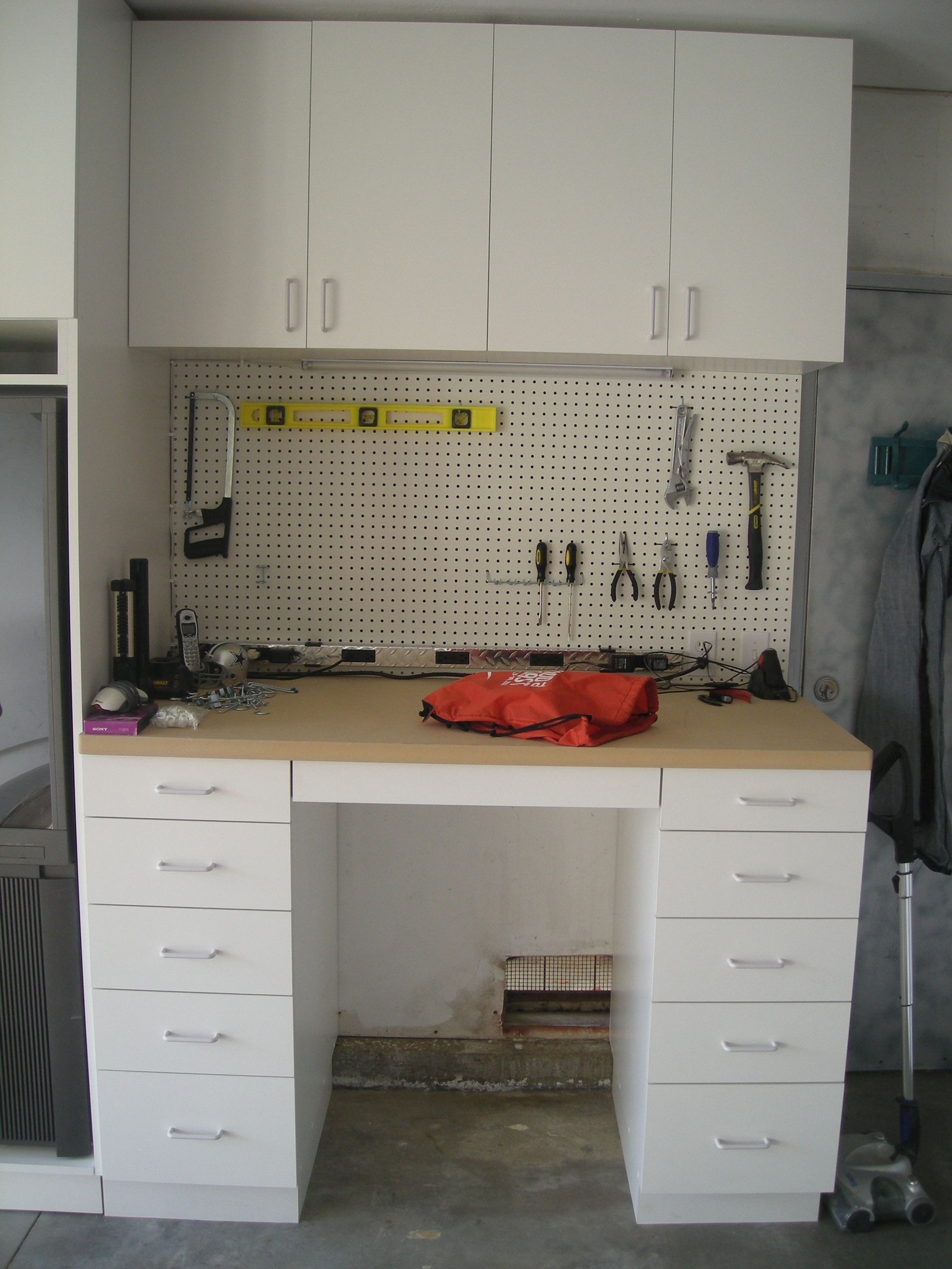 White workbench with drawers and overhead cabinets, tools on pegboard, in garage.