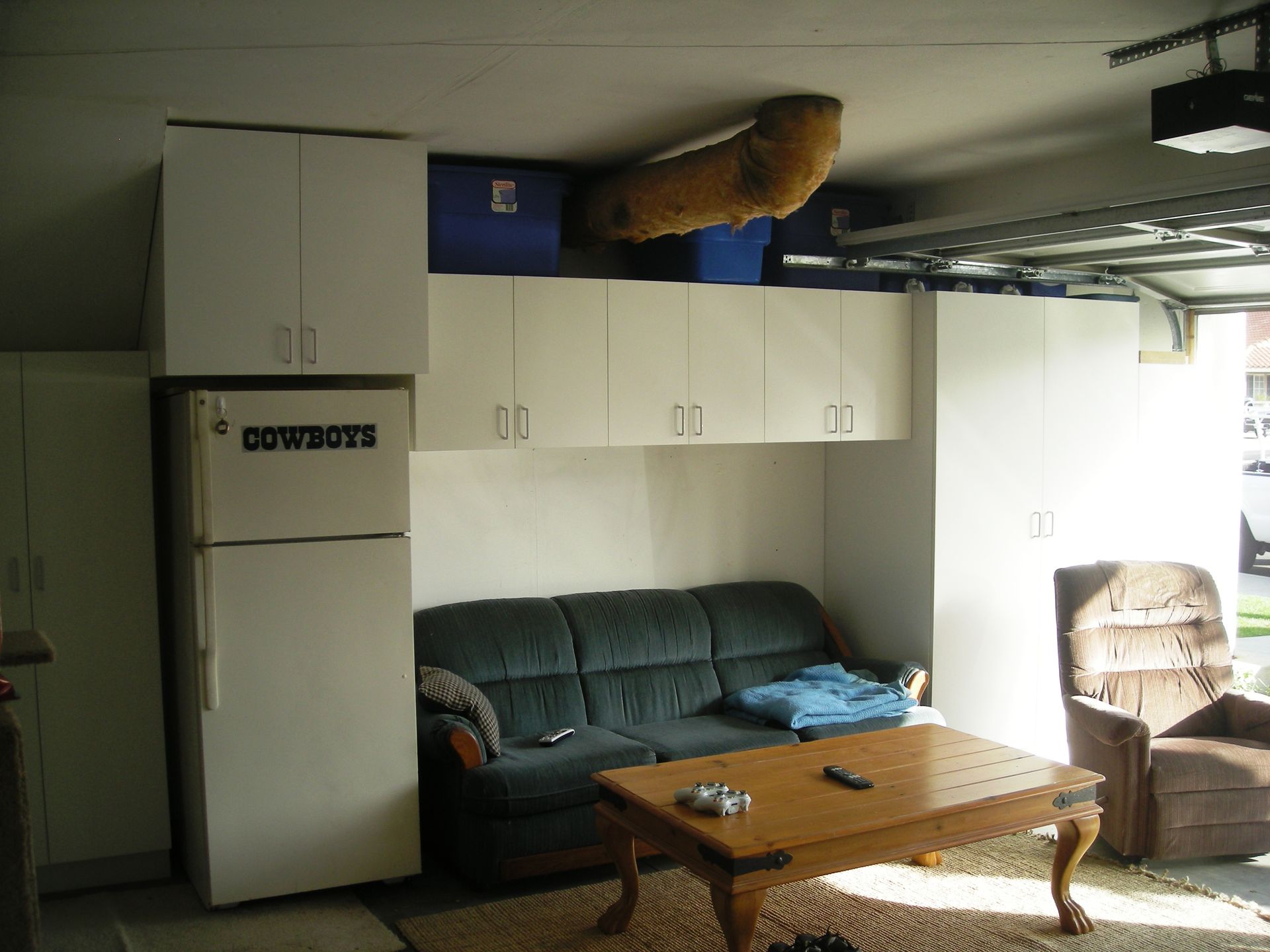 Garage with a couch, cabinets, refrigerator, and a table. A large object is mounted on the ceiling.