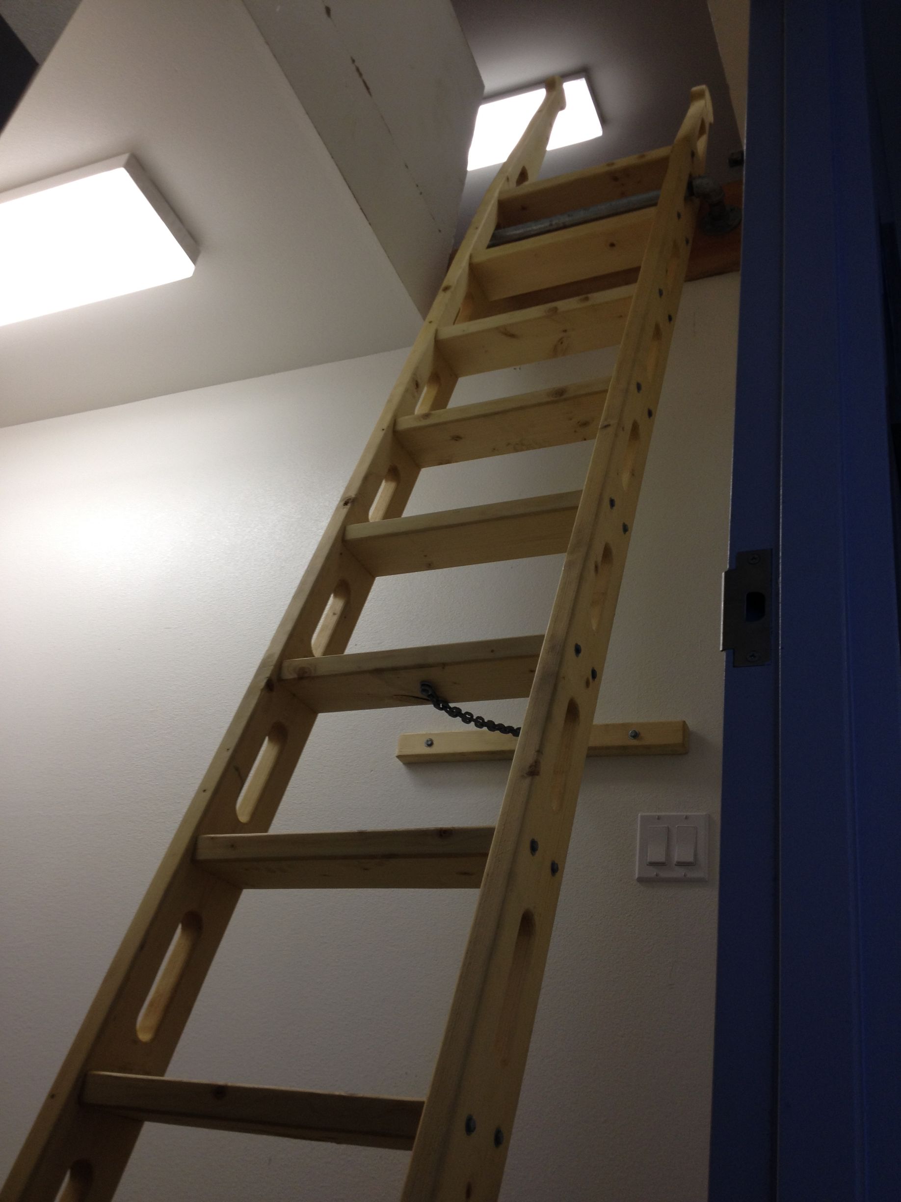 Wooden ladder leading to an attic entrance. Light-colored wood, attached to a white wall.