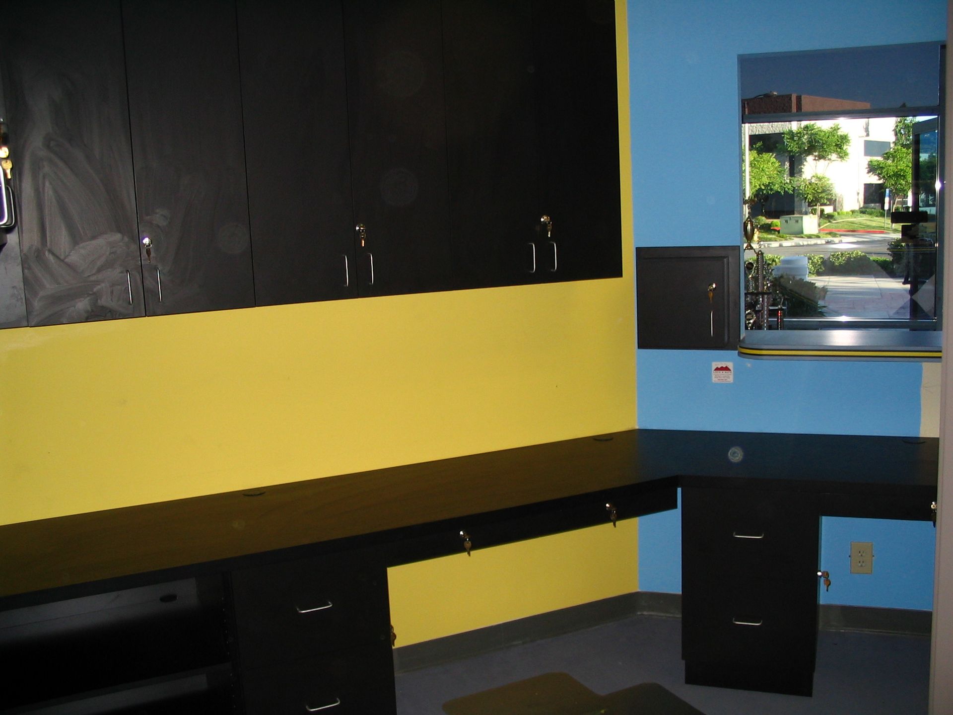 Black workspace with yellow and blue walls, cabinets, and a window with an outdoor view.