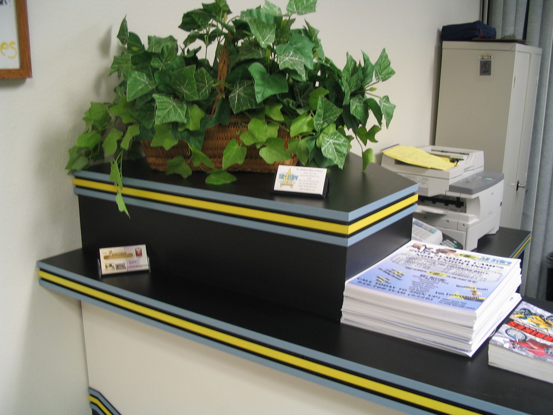 Plant arrangement atop a black tiered counter with yellow and blue trim; printer and stacked publications nearby.
