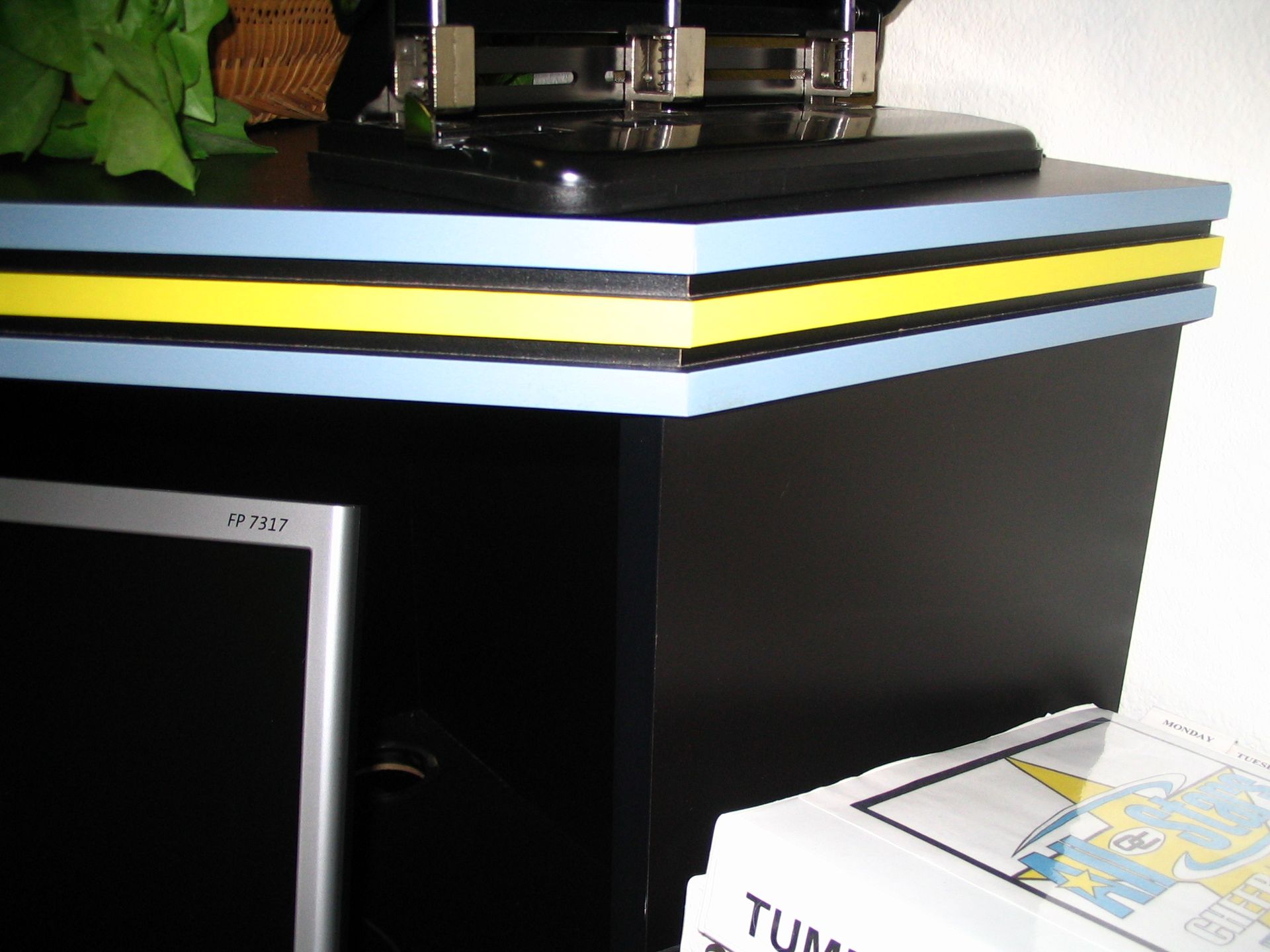 Black cabinet with a blue and yellow trimmed edge, a monitor, and a stapler.