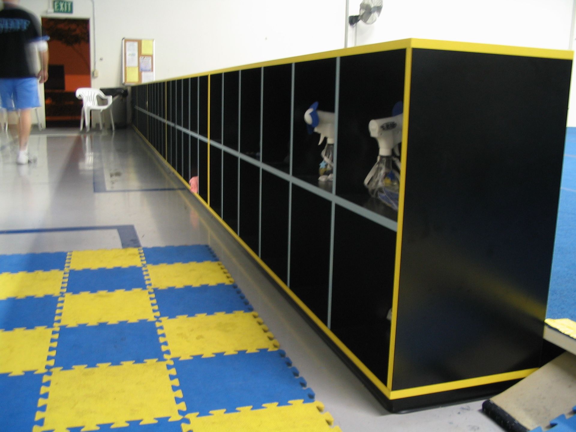 Long row of black lockers with yellow trim in a gymnasium, blue and yellow mat in foreground, person walking in background.