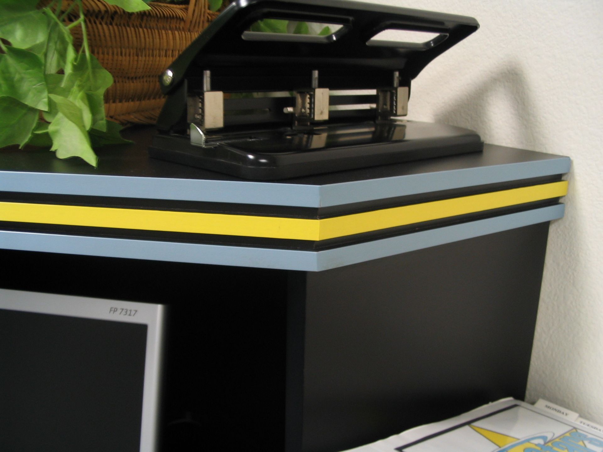 Black desk with blue and yellow trim, a hole punch, and a computer monitor.