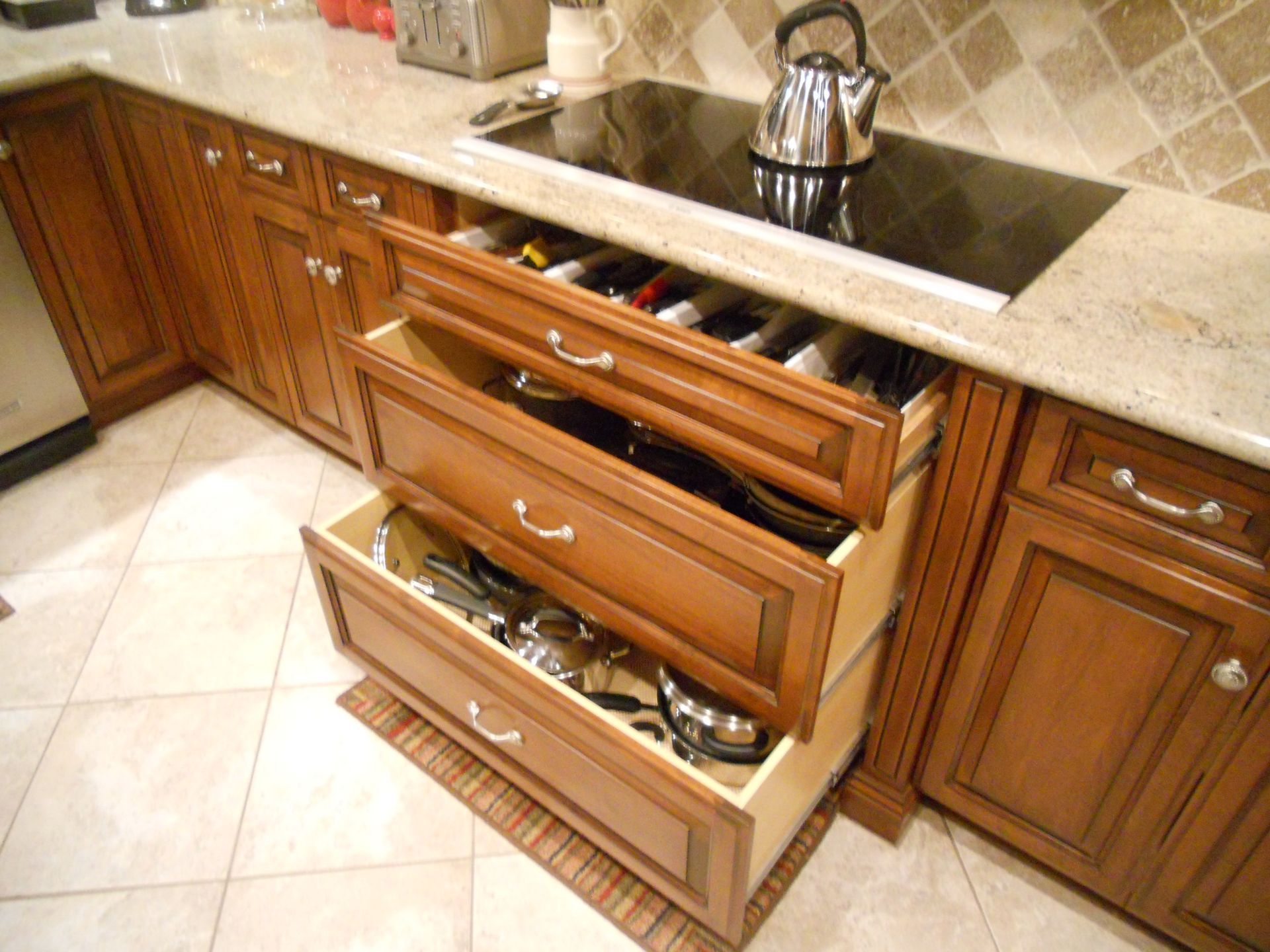 Kitchen drawers pulled out, showing utensils, pots, and pans, beneath a stovetop and countertop.