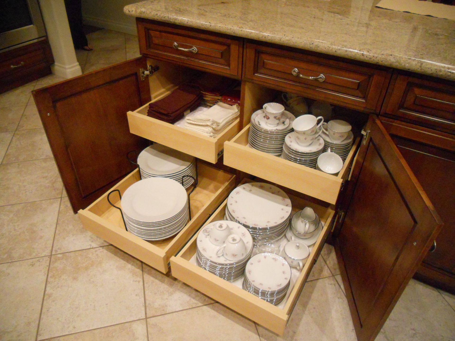 Kitchen cabinet with pull-out drawers holding plates and teacups. Dark wood cabinetry; light-colored stone countertop.