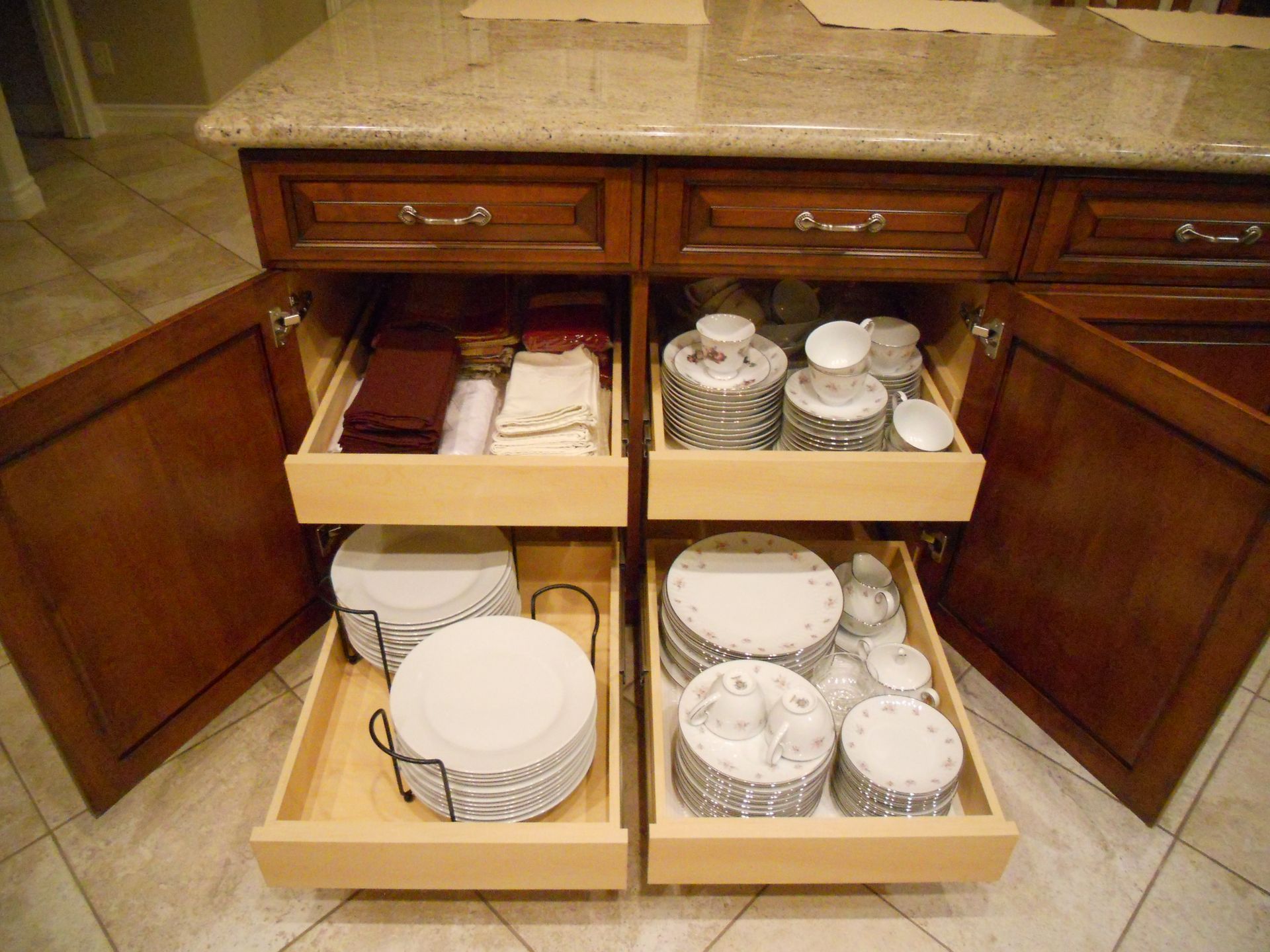 Kitchen cabinet with pull-out drawers holding dishes and linens. Wooden cabinets and a granite countertop.