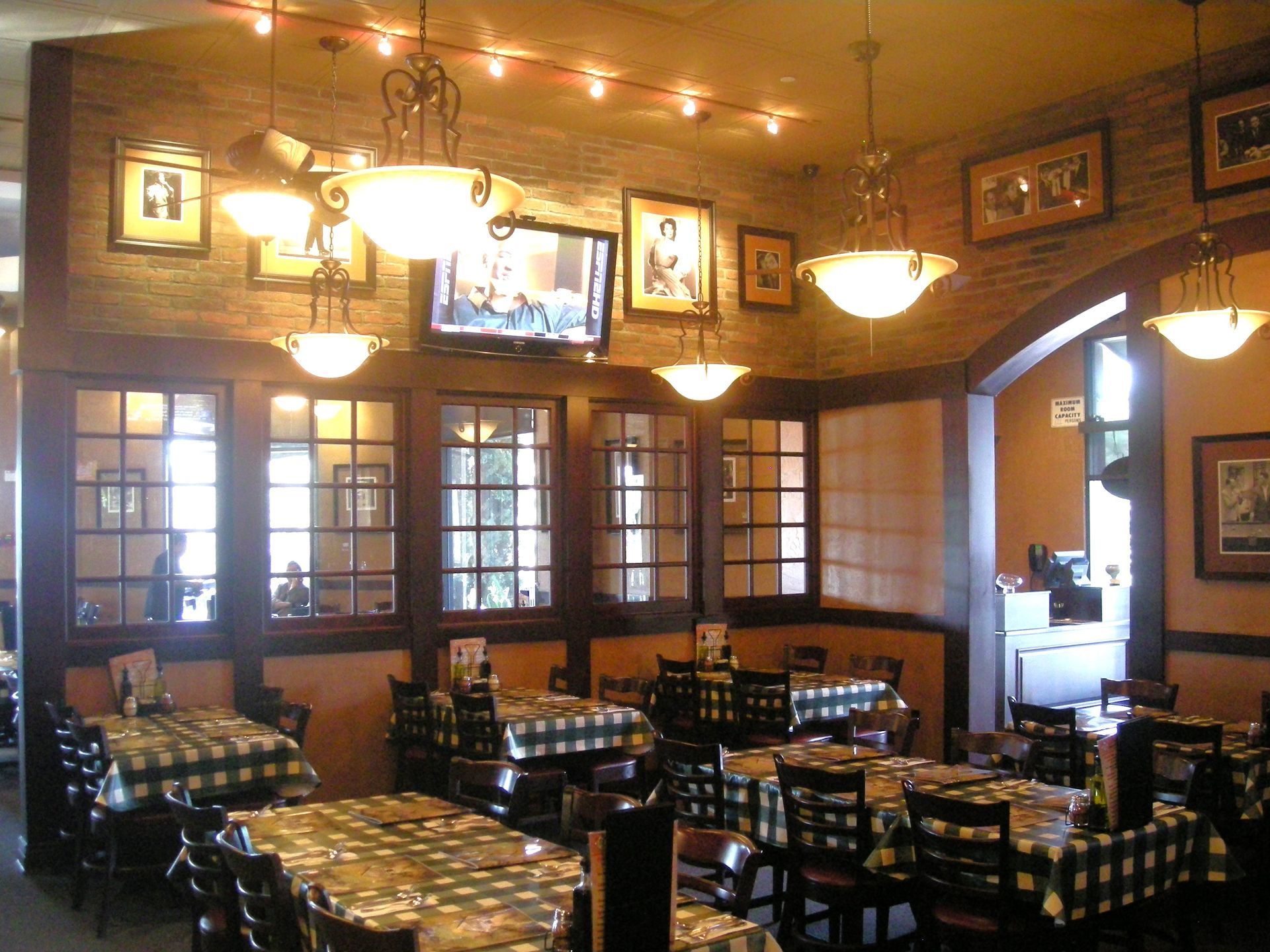 Restaurant interior with tables, checkered tablecloths, windows, brick wall, and hanging lights.