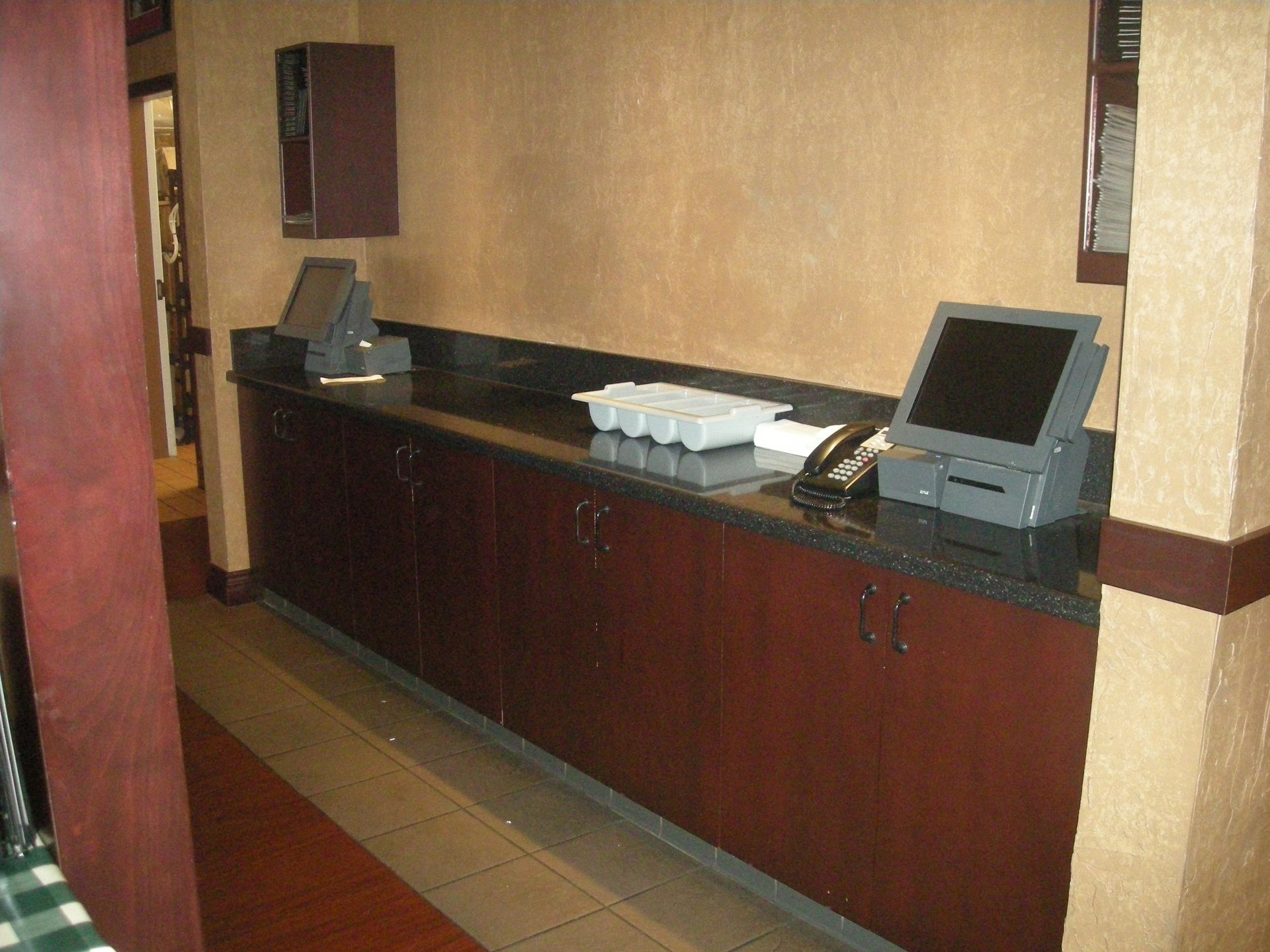 Dark wood counter with granite top, two computer terminals, phone, and serving supplies in a restaurant.