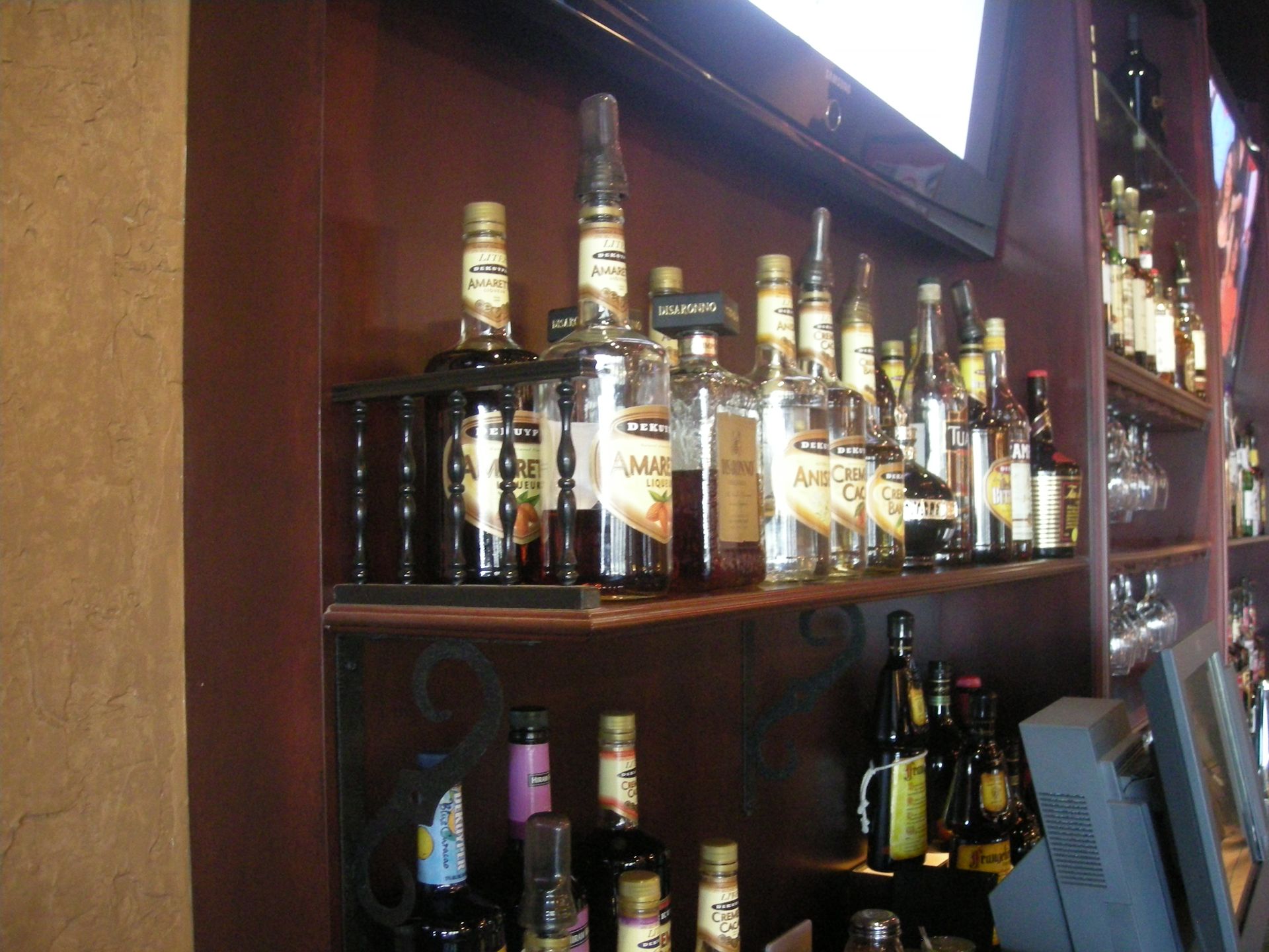 Bar shelf with various liquor bottles, brown wood, some with gold labels, lit from above.
