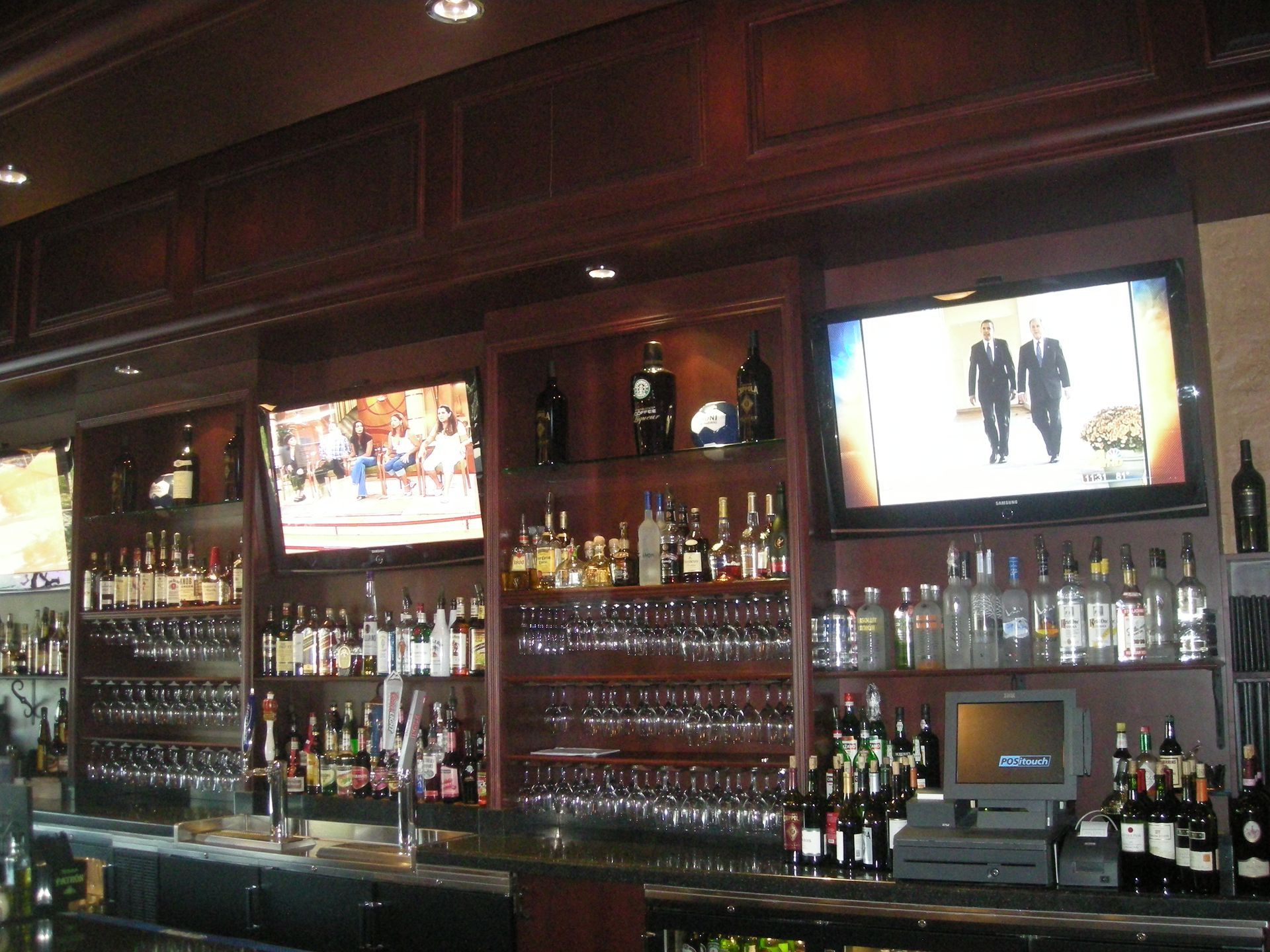 Bar with dark wood shelves filled with bottles, glasses, and three TVs showing different content.