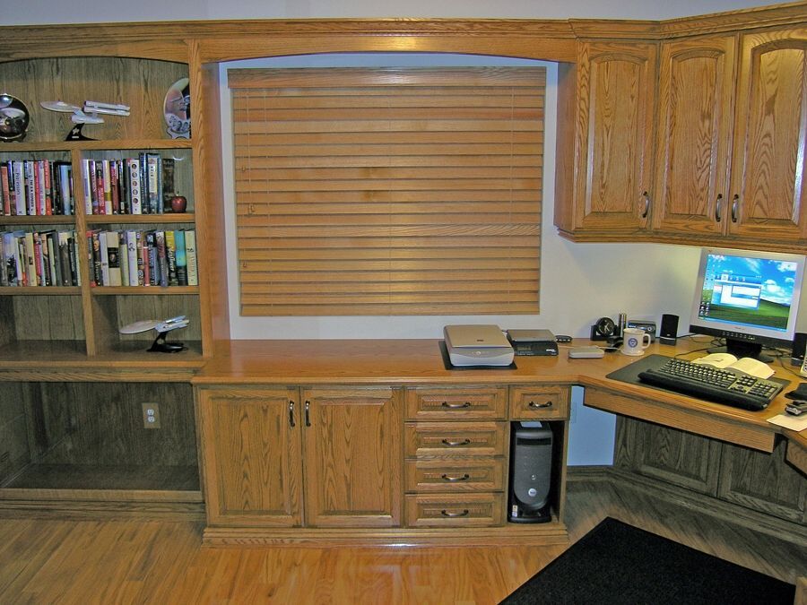 Built-in oak home office with bookshelves, desk, cabinets, computer, and a slatted window treatment.