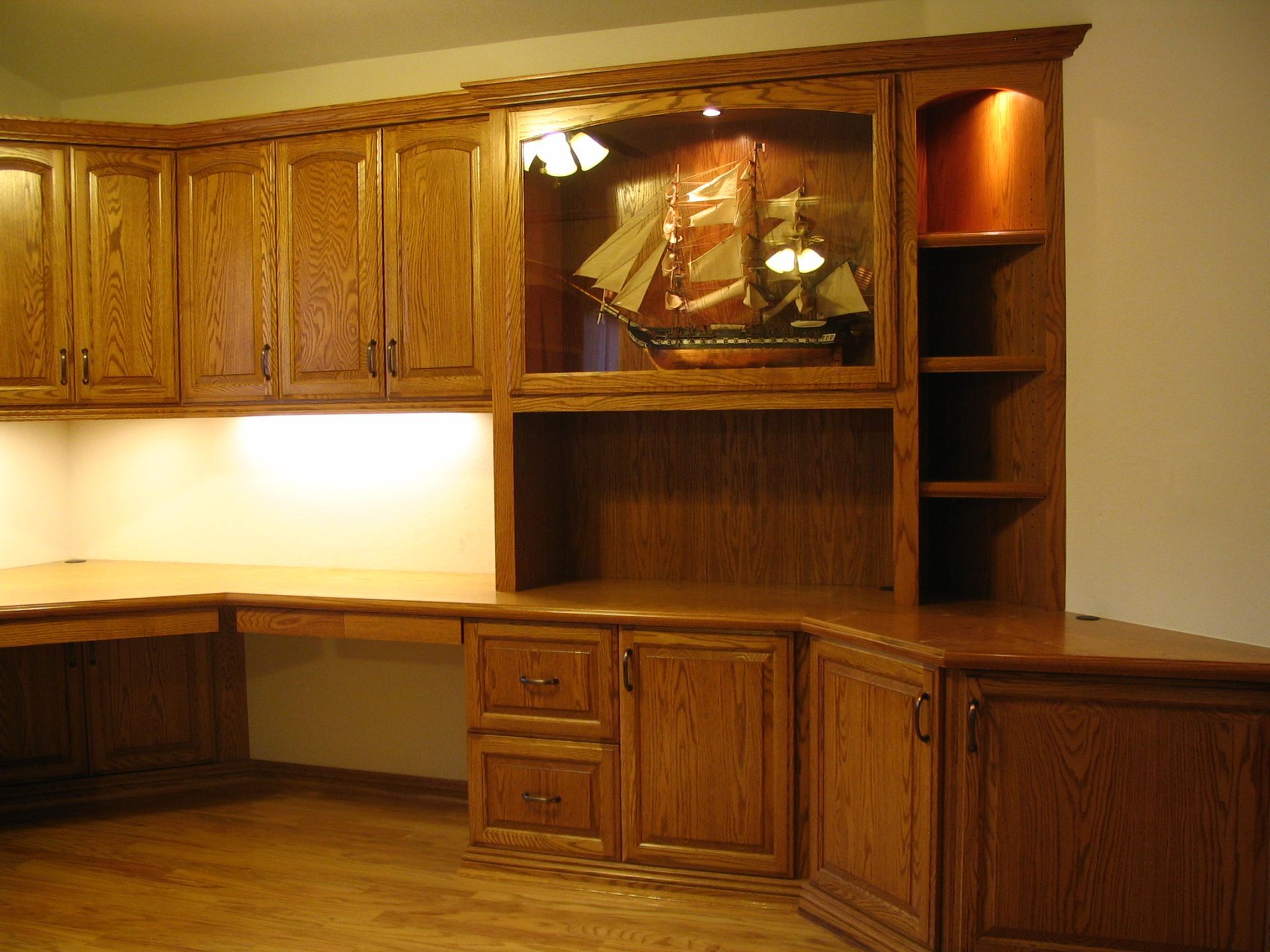 Wooden built-in desk and cabinets with a display shelf holding a ship model; warm lighting.