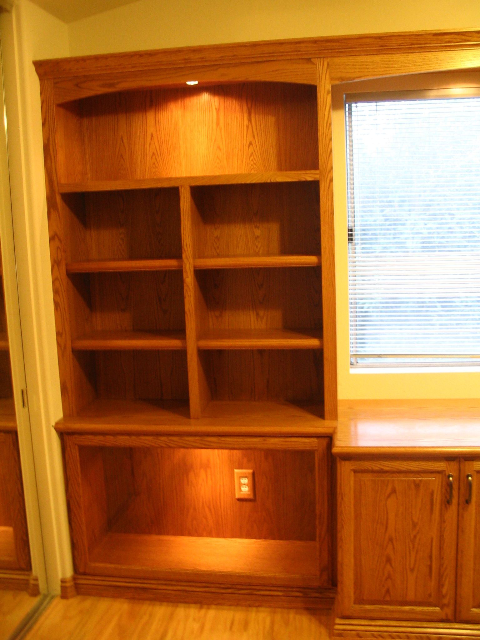 Built-in wooden bookcase with recessed lighting, next to a window and cabinet, all with a warm, reddish-brown stain.