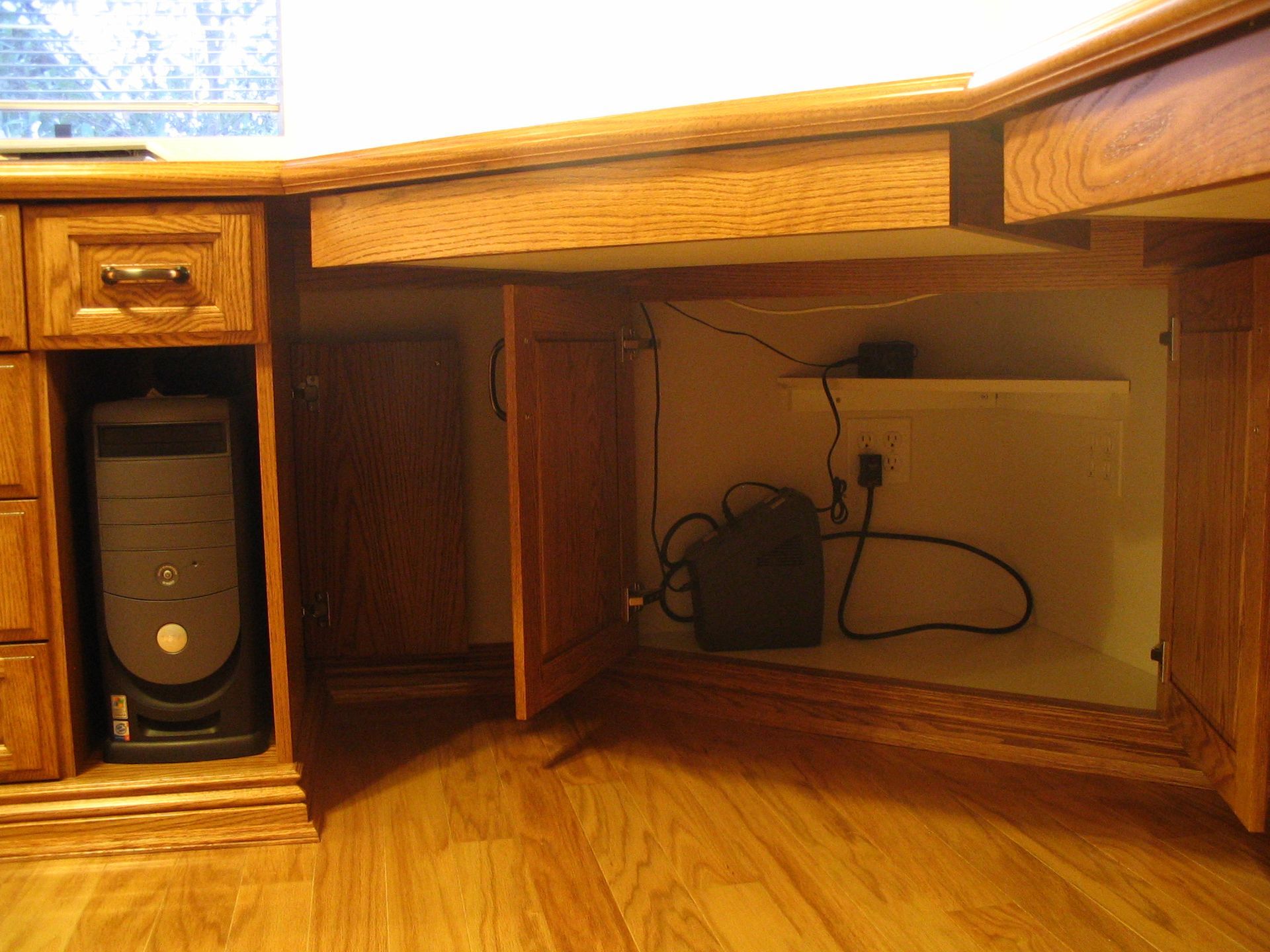 Computer tower inside desk cabinet, with power supply and cords. Wooden desk, cabinets, and floor.