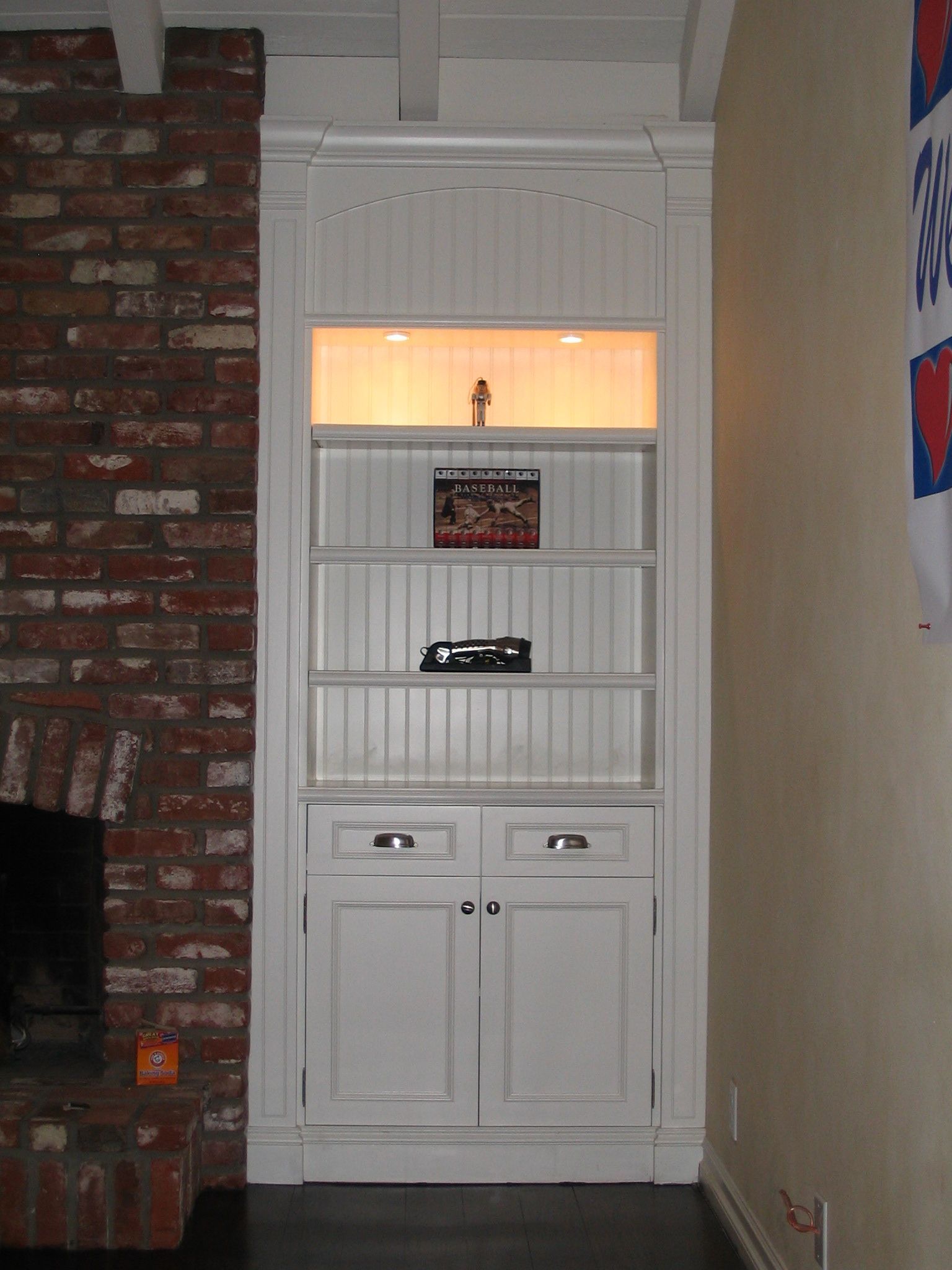 Built-in white cabinet in an alcove with shelves and doors, lit interior. Next to a brick fireplace.