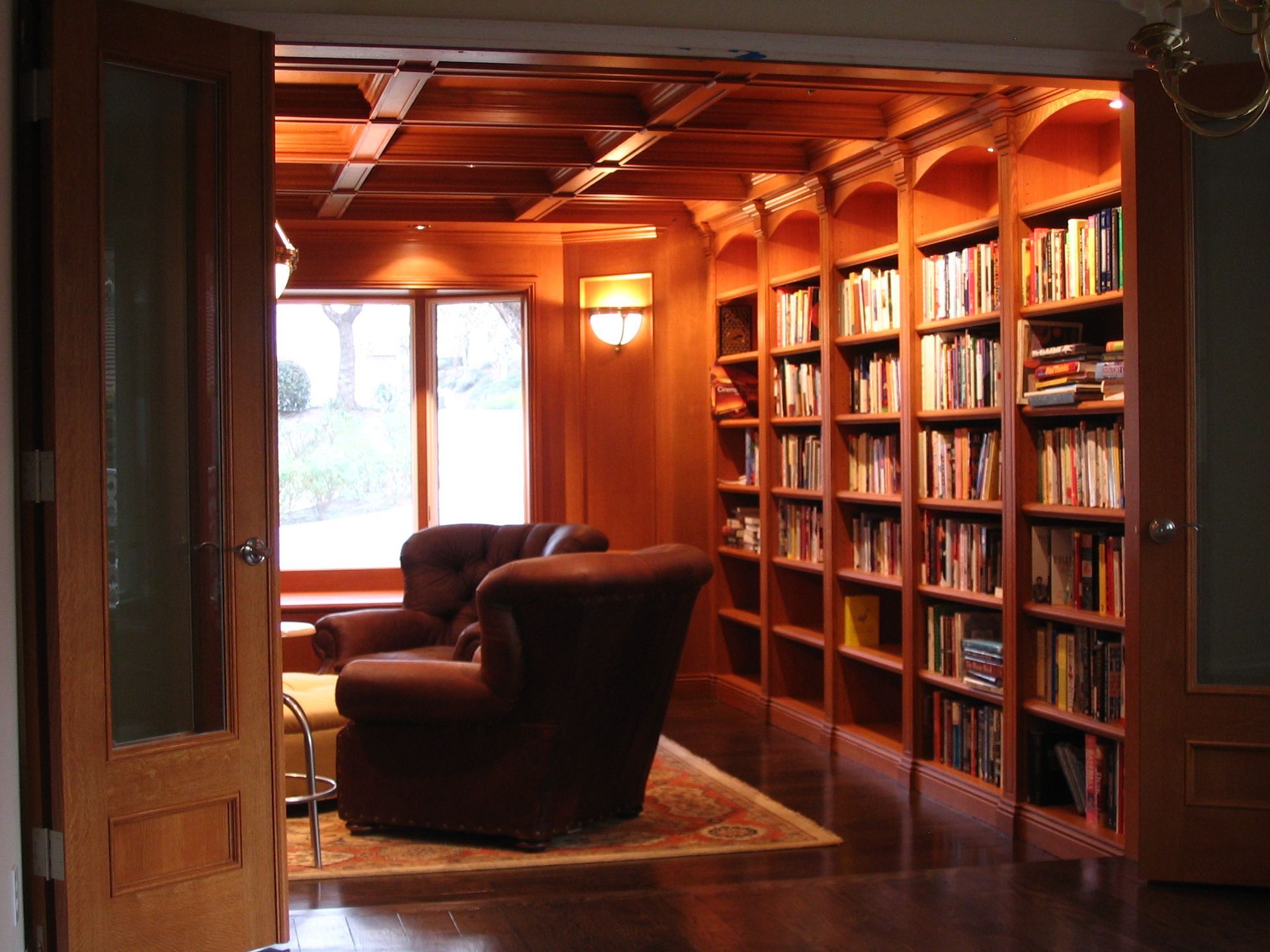 Cozy library with built-in bookshelves, brown armchairs, wooden ceiling, and natural light from a window.