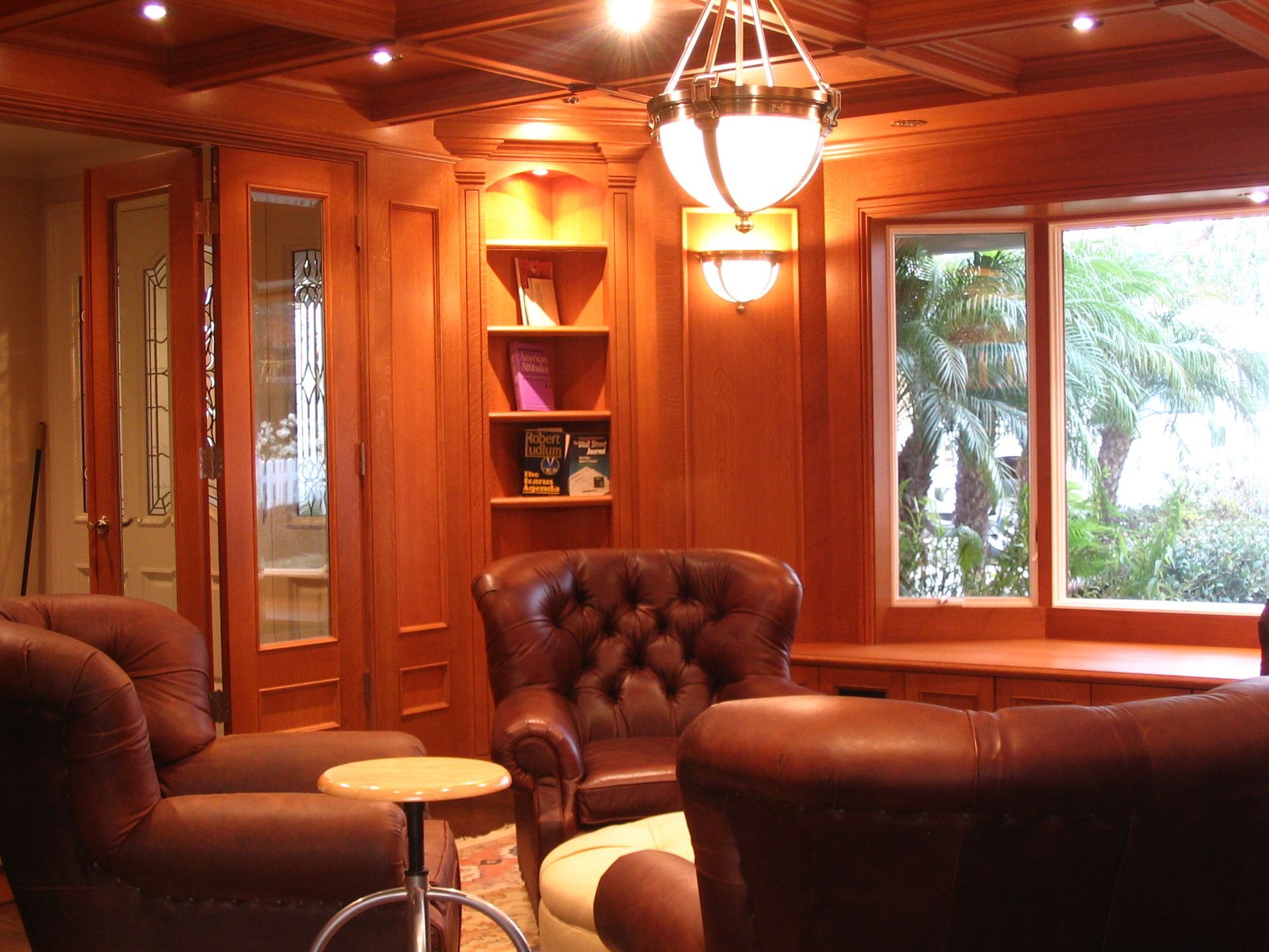 Cozy library with leather armchairs, built-in bookshelves, and a window. Wooden paneling and ornate ceiling with lighting.
