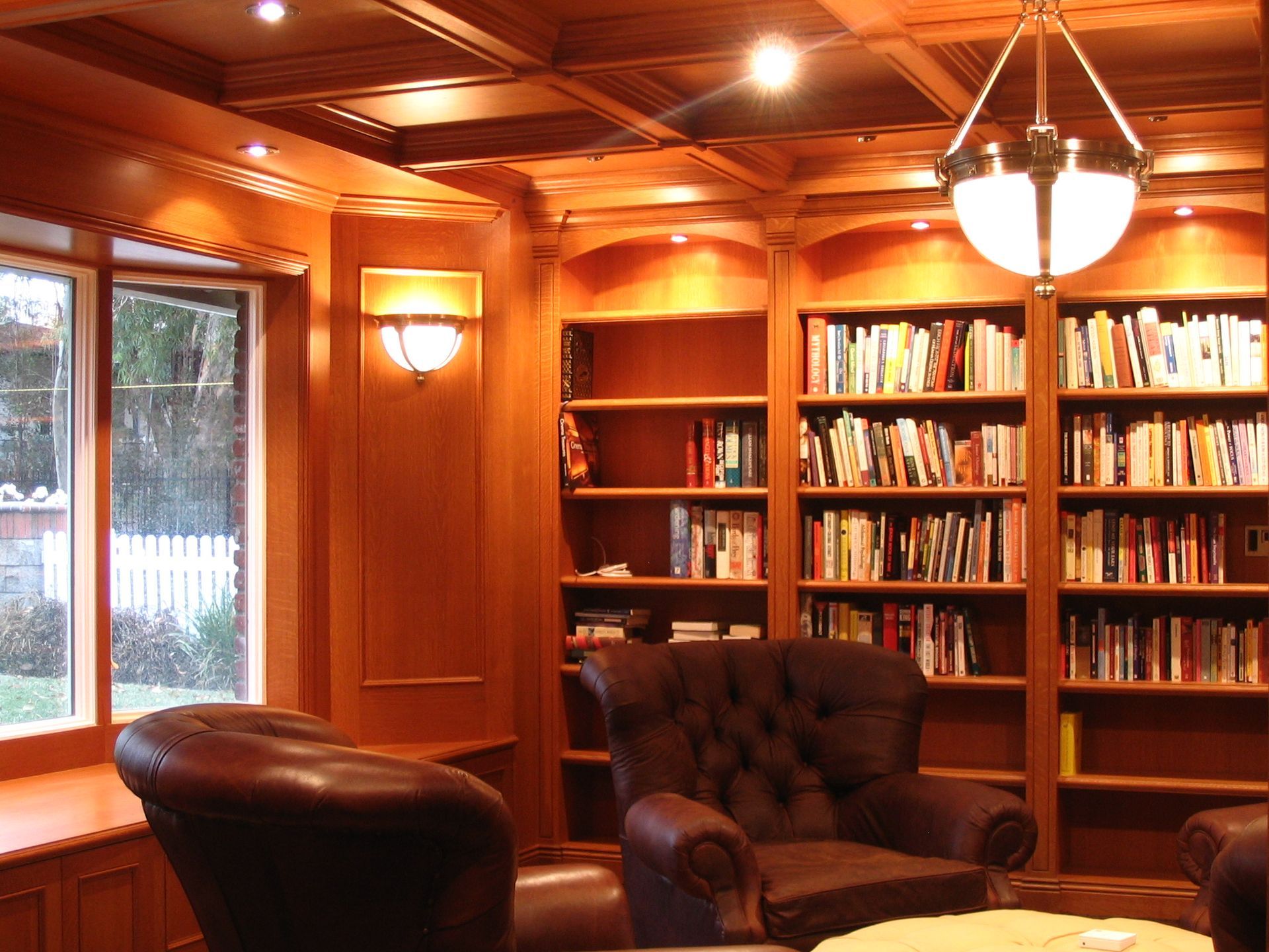 Cozy wooden library with built-in bookshelves, leather armchairs, bay window, and soft lighting.