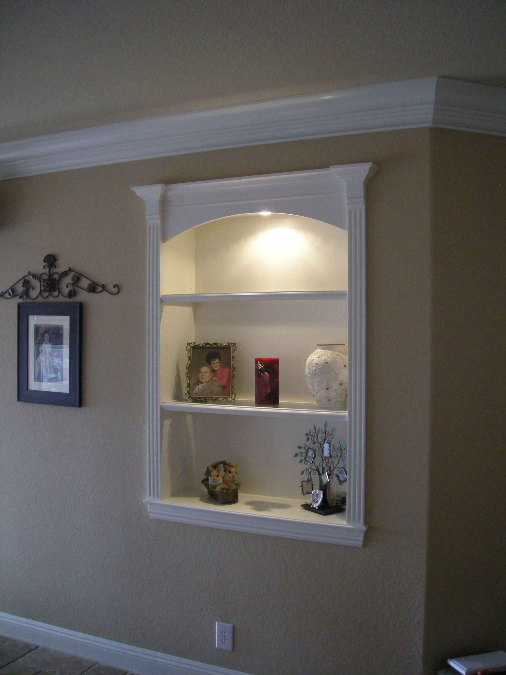 Built-in white shelf niche with arched top, lit display shelves, decorated with items on a beige wall.