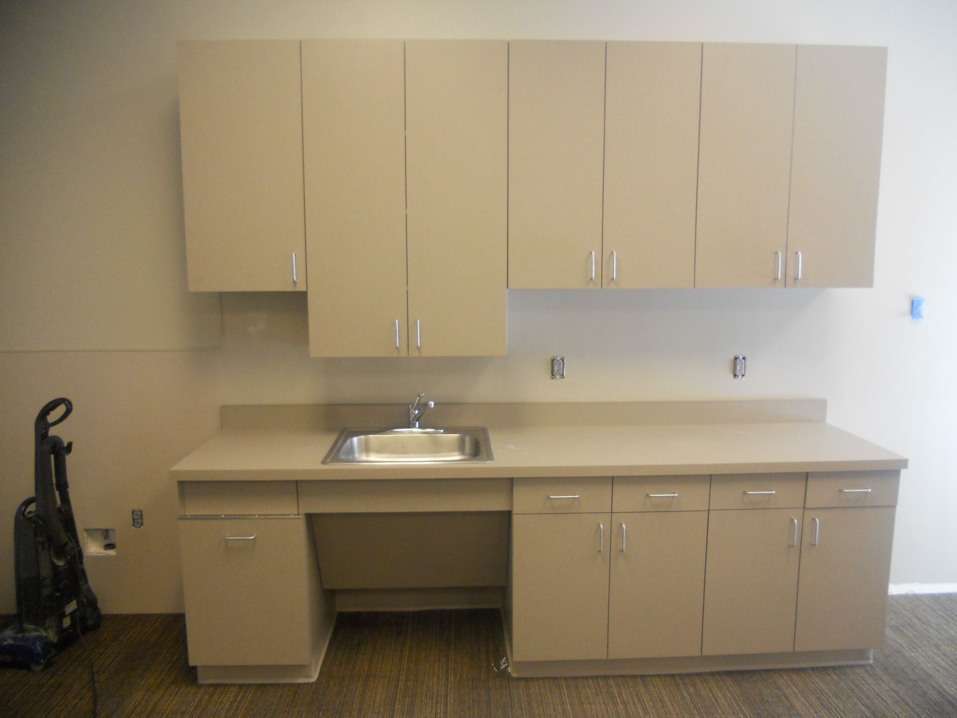 Beige cabinetry with a sink, countertop, and vacuum cleaner in a room.