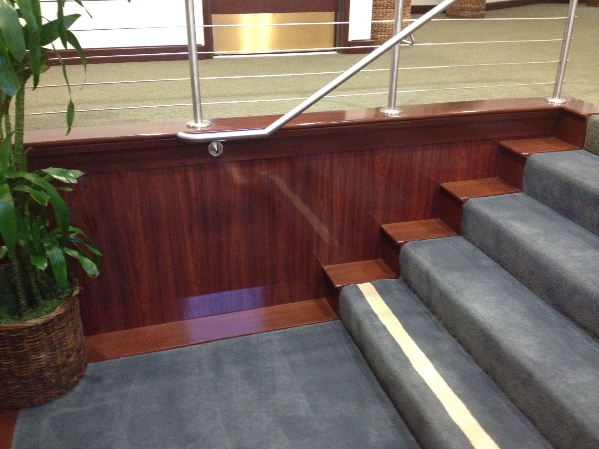 Staircase with gray carpeted steps, dark wood paneling, and a metal handrail, indoors. A potted plant is on the side.