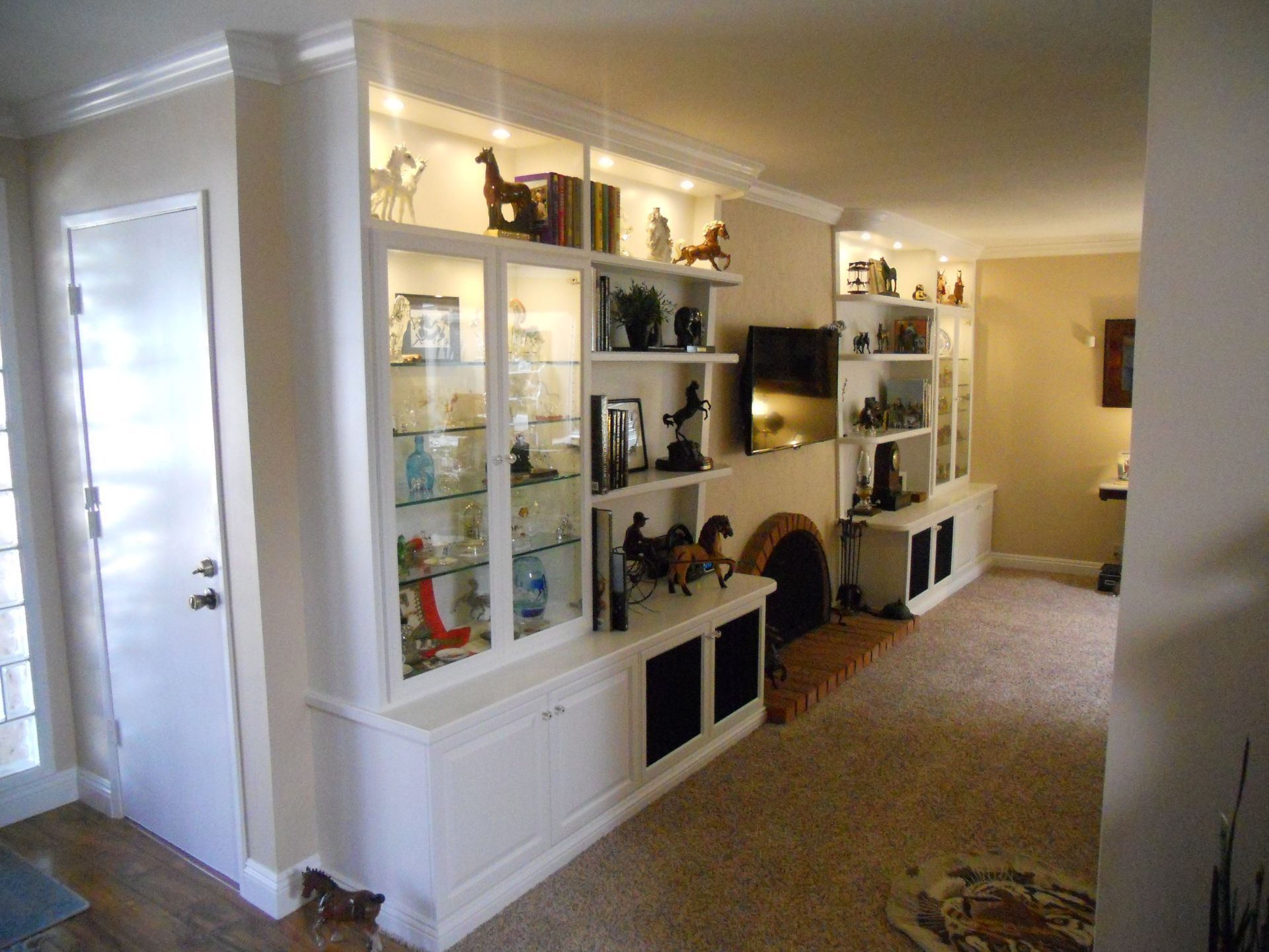Built-in white cabinets with glass doors, shelves, and lighting in a living room, alongside a fireplace.
