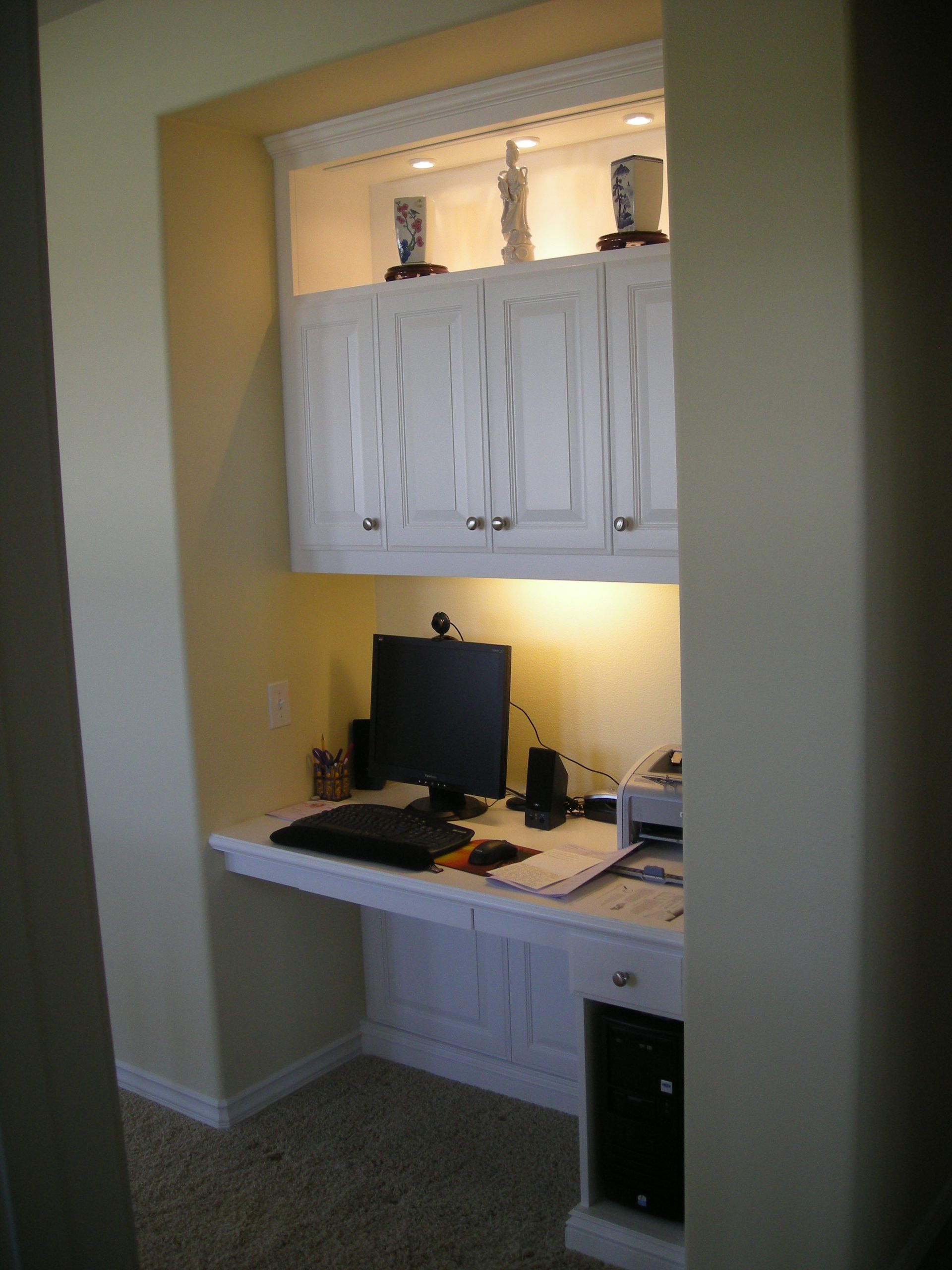 Built-in white desk and cabinets in a small alcove, with a computer and office supplies; lit from above.